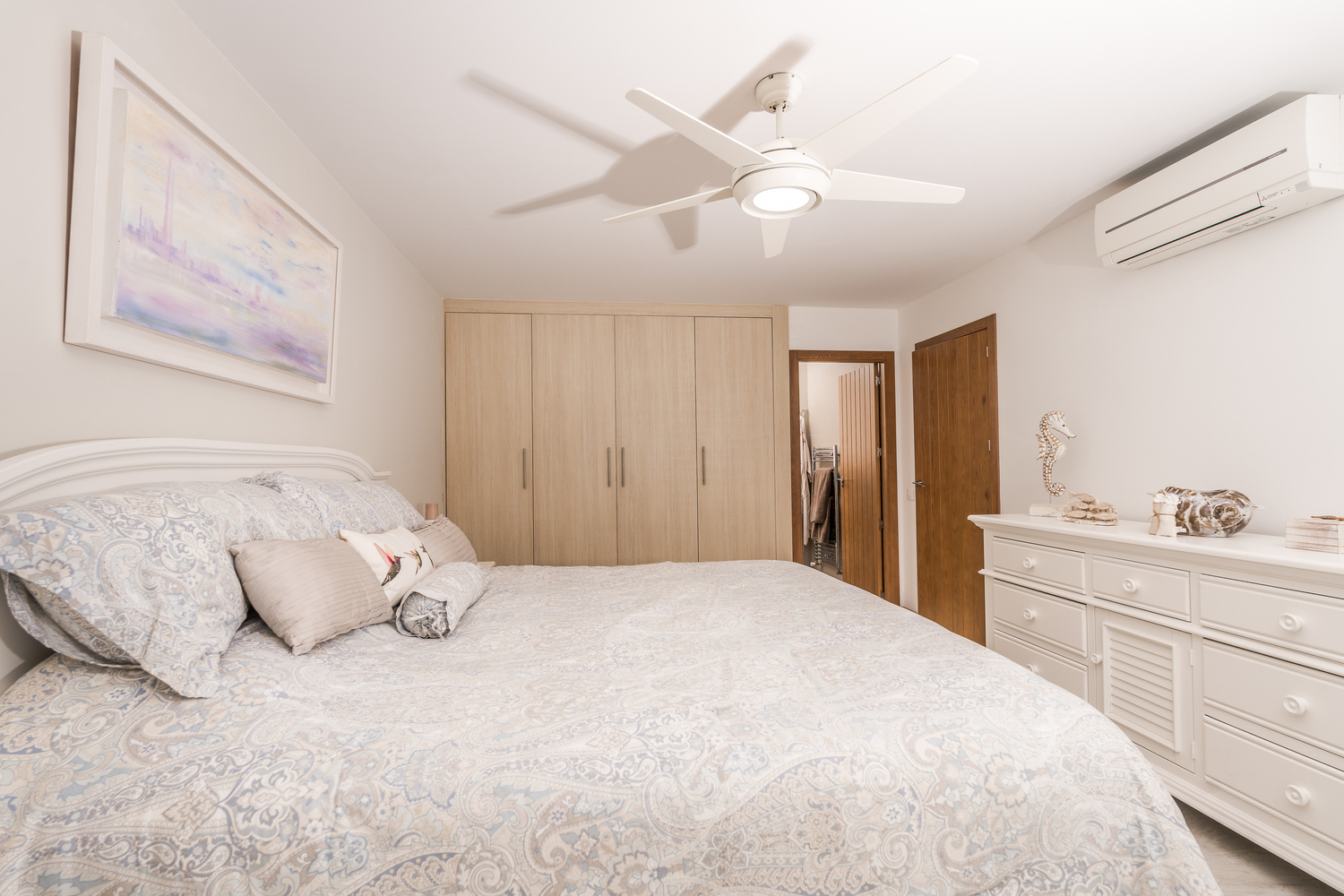 Modern bedroom featuring built-in light wood wardrobes, a white dresser, and a ceiling fan. Includes wall-mounted air conditioning and an en-suite access point.