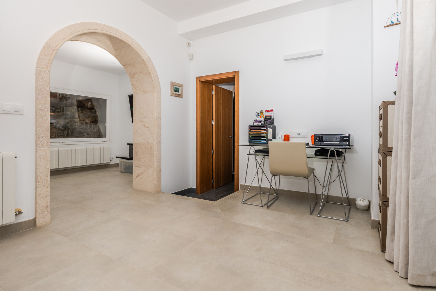 Office area featuring a glass desk and a traditional Tosca stone archway leading to an adjacent room with neutral tiled flooring.