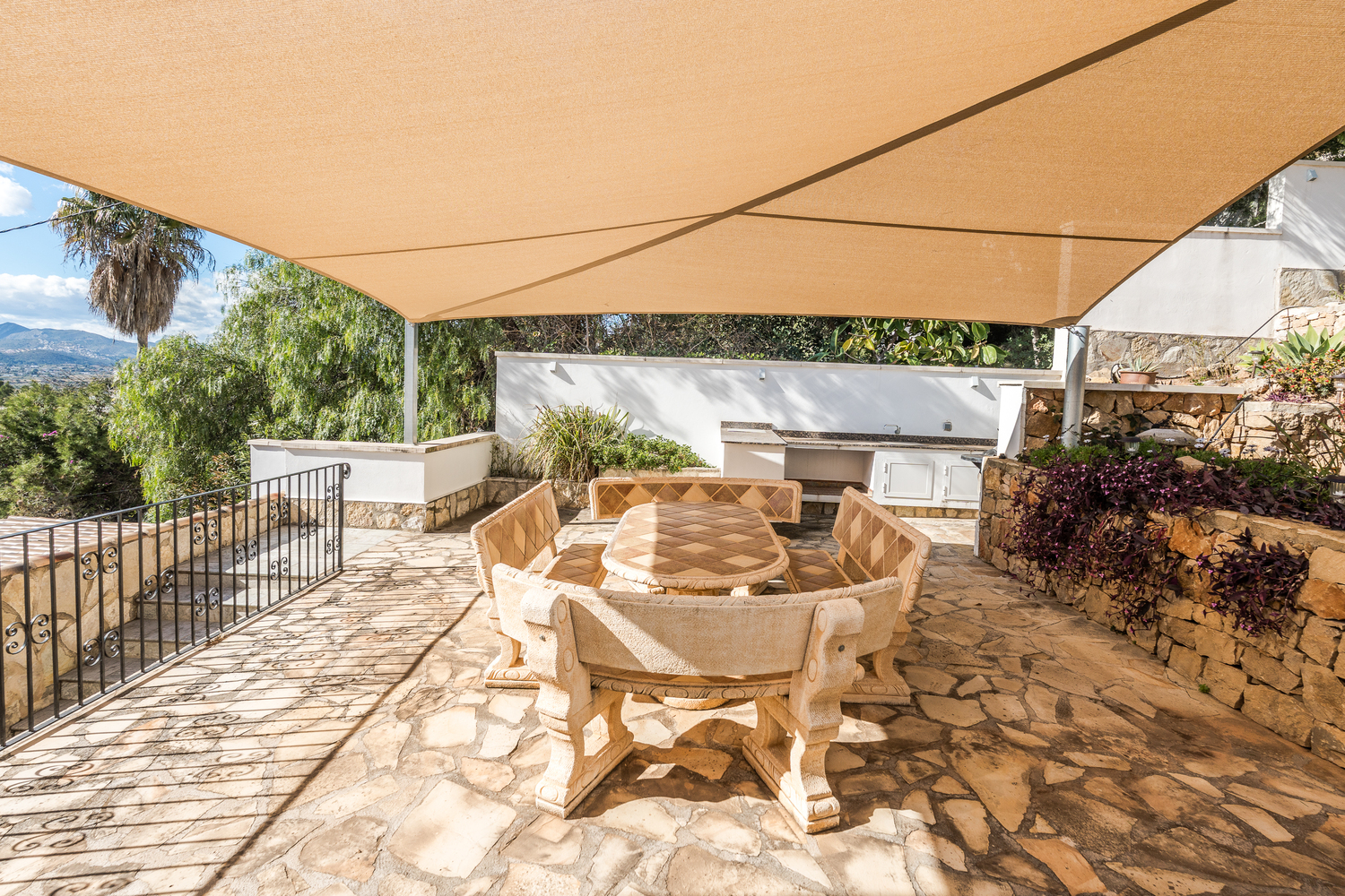 Outdoor dining area featuring a carved stone table and benches, shaded by a large beige sun sail on a flagstone terrace adjacent to the summer kitchen.