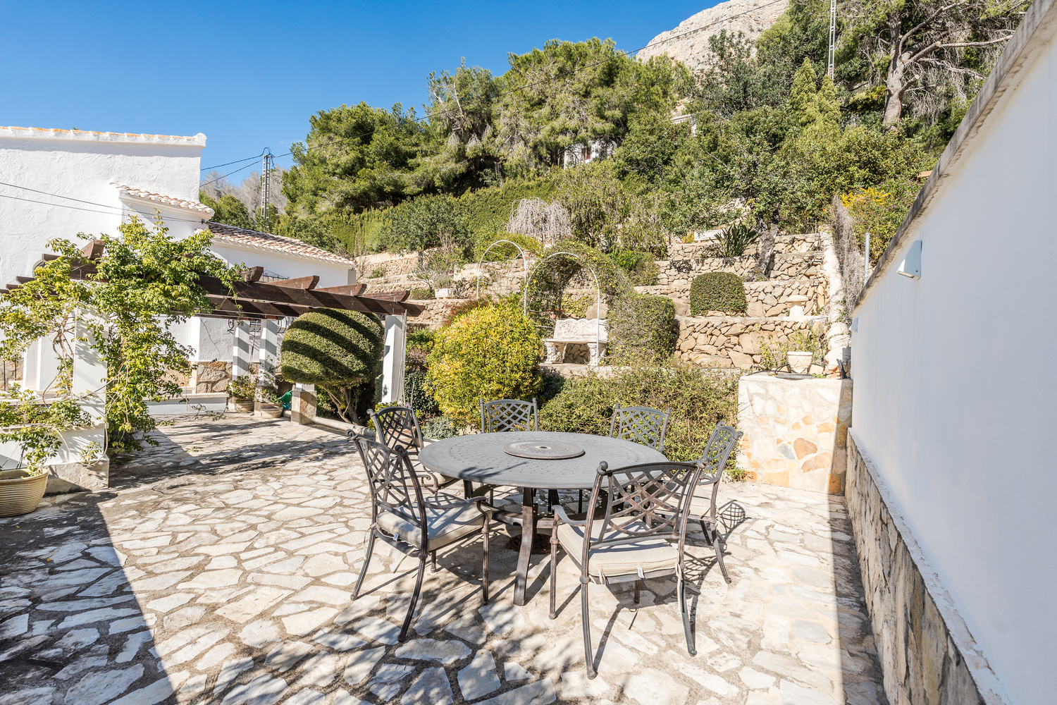 Outdoor dining area featuring a round wrought-iron table on a natural stone patio, set against tiered Mediterranean gardens and a wooden pergola.