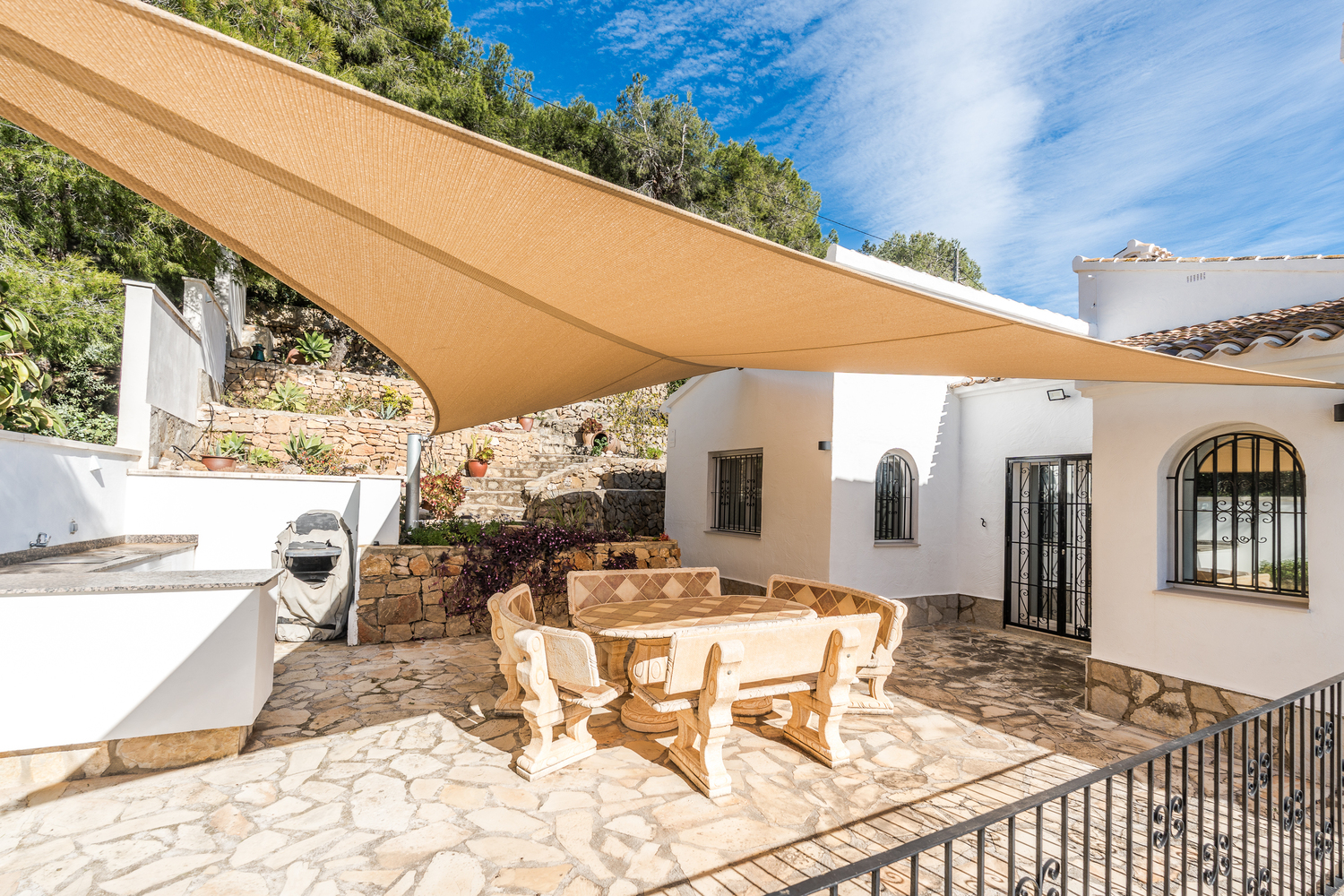 Outdoor dining terrace featuring a stone table and bench set under a large sun sail, surrounded by natural stone walls and Mediterranean landscaping.