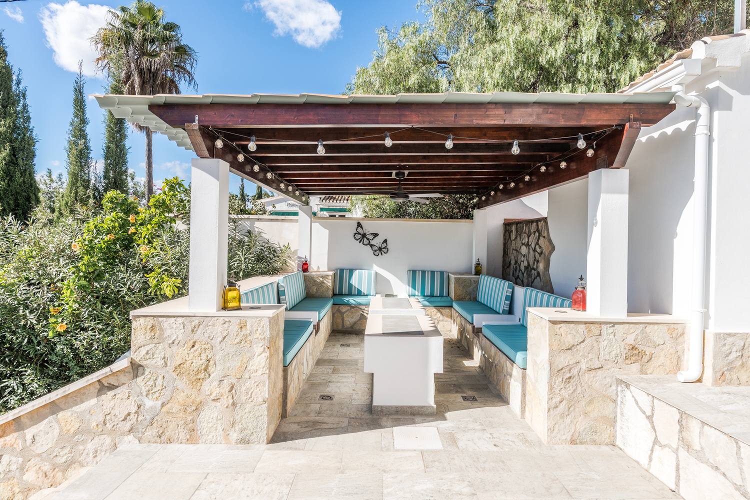 Outdoor lounge area featuring a wooden pergola with string lights, a ceiling fan, and built-in stone seating with turquoise striped cushions.
