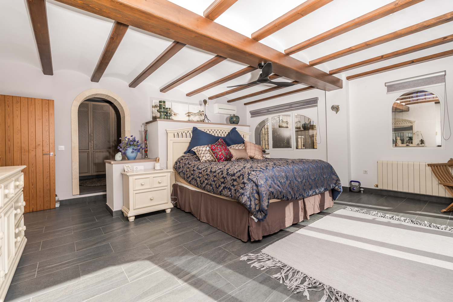 Spacious bedroom featuring exposed wooden beams, grey slate flooring, and a stone-arched entrance leading to a dressing area. Includes air conditioning and central heating.