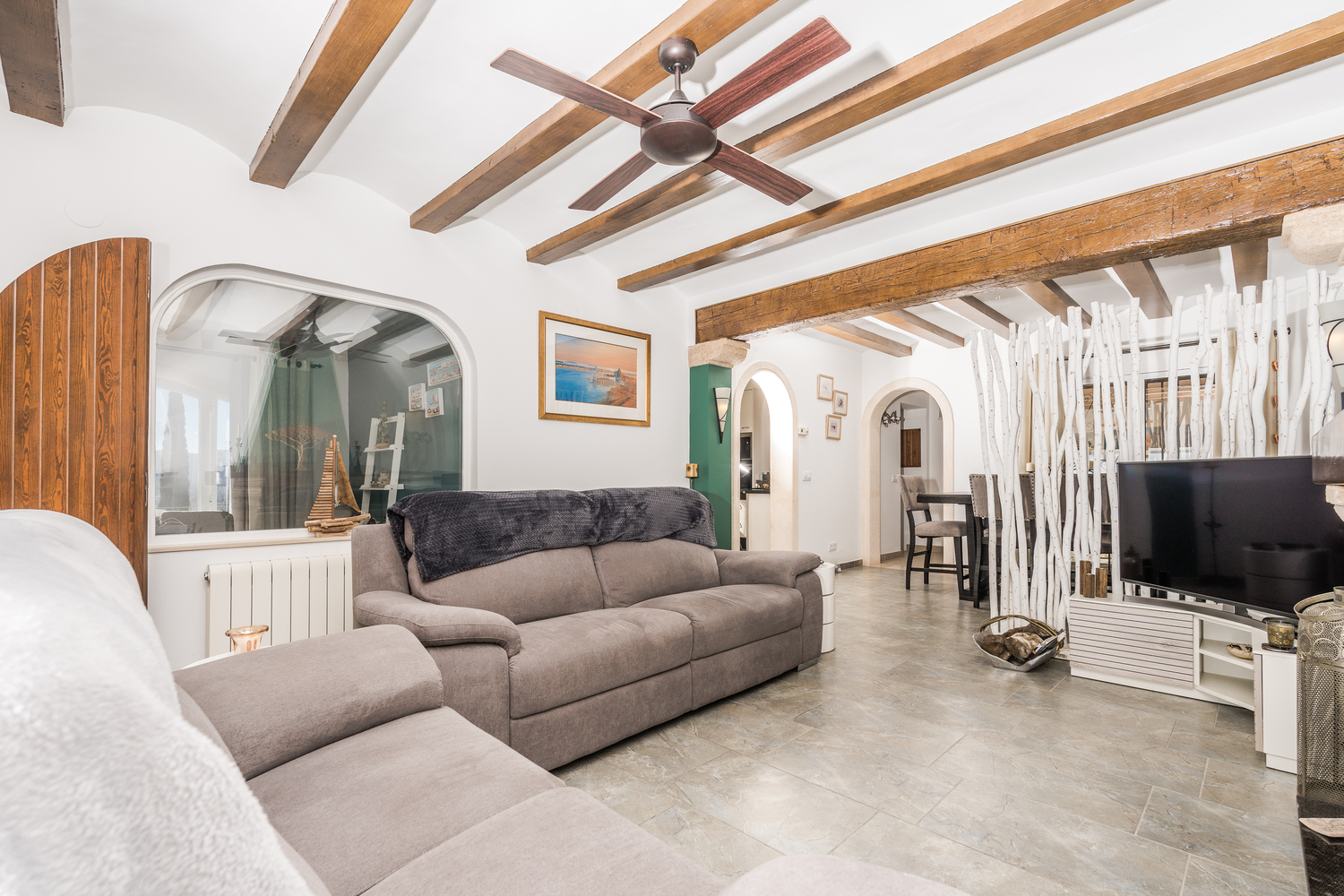 Spacious living area featuring rustic wooden ceiling beams, a ceiling fan, and tiled flooring. A decorative branch screen separates the lounge from the dining area.
