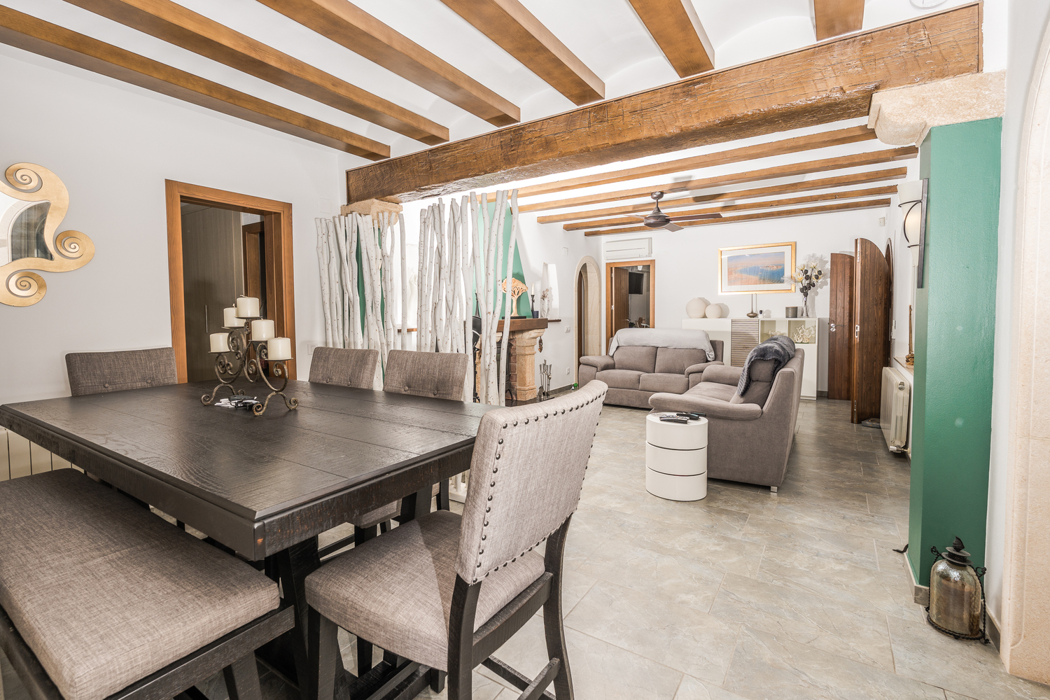 Spacious living-dining area featuring exposed wooden ceiling beams, stone tile flooring, and a large dark wood table with upholstered seating.