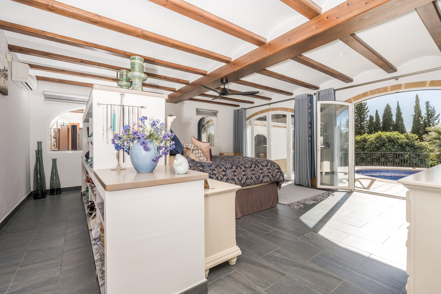 Spacious master bedroom featuring exposed wooden ceiling beams, dark tiled floors, and arched glass doors providing direct access to the private swimming pool.