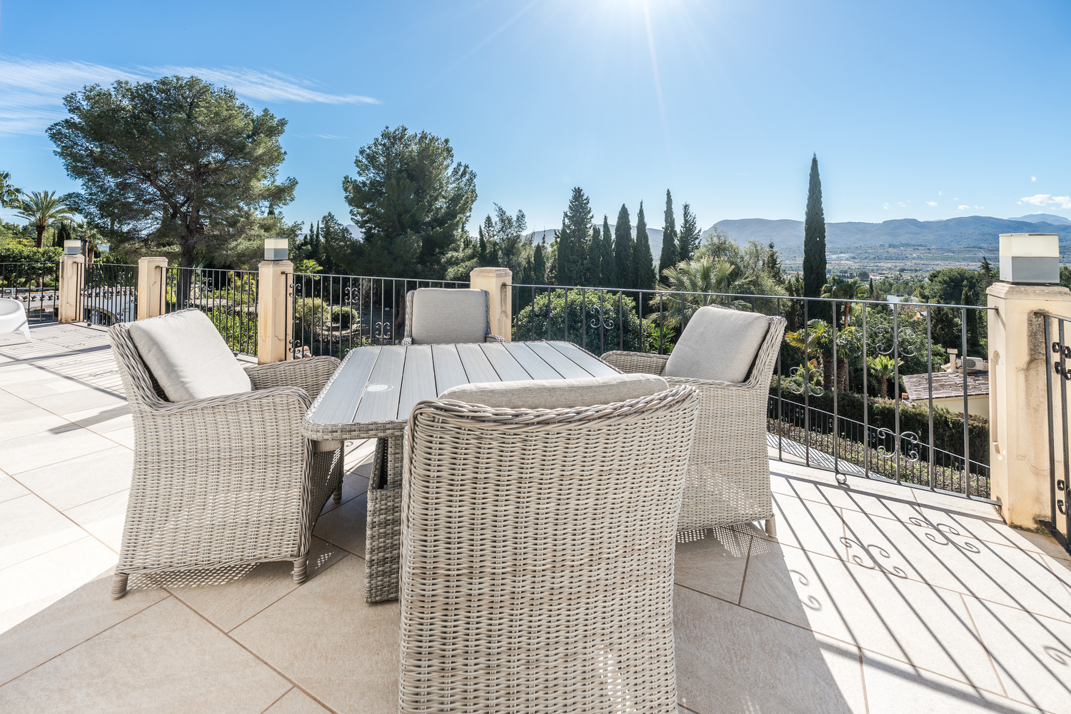 Spacious outdoor terrace featuring a wicker dining set and panoramic valley views. The area is finished with light stone tiling and a wrought iron railing.