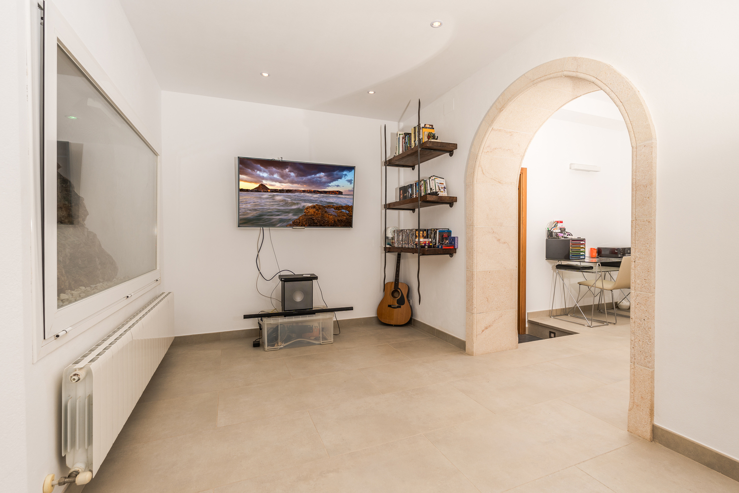 TV room featuring a wall-mounted screen, rustic wooden shelving, and a natural stone archway leading to a dedicated office space with a desk.