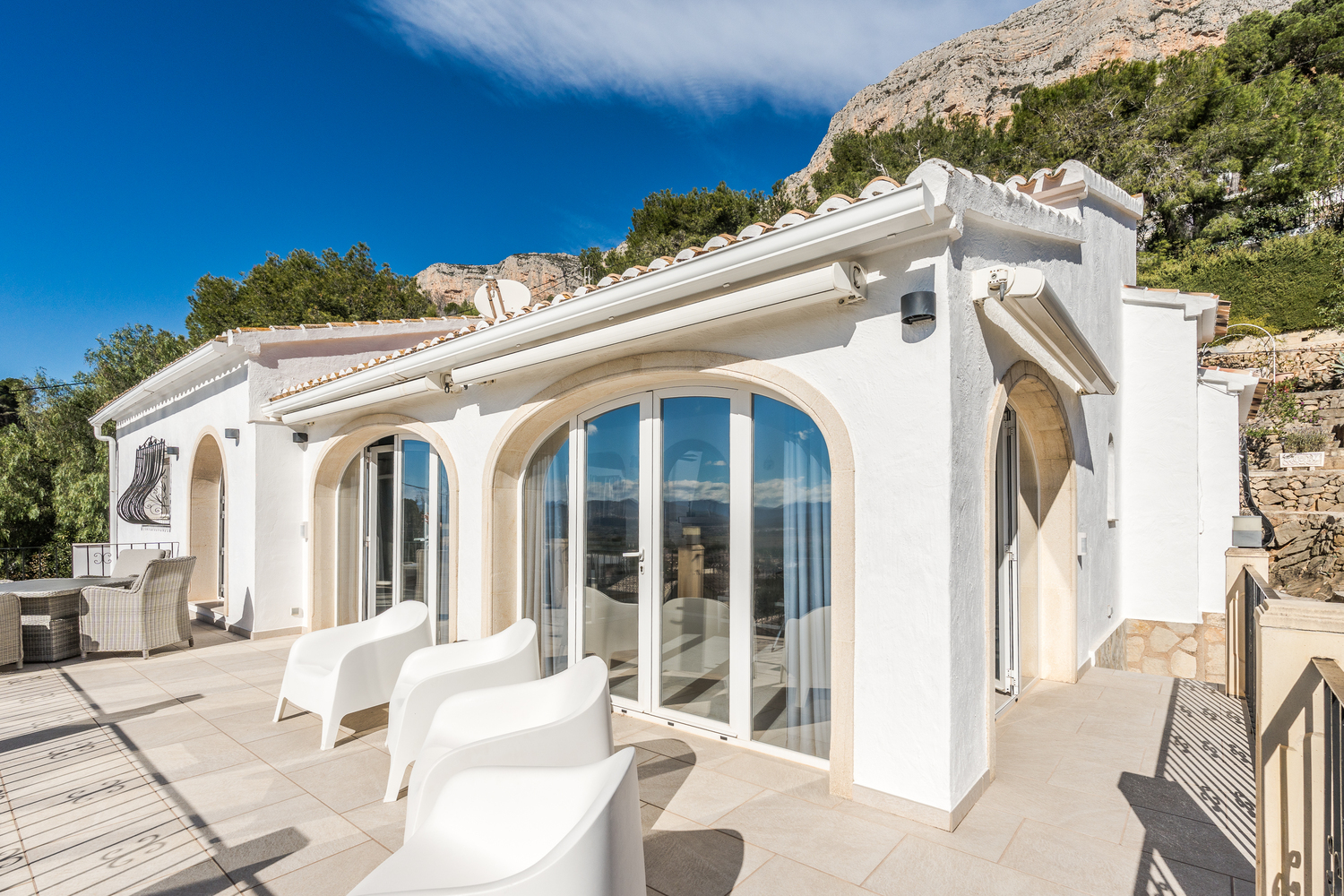 Upper terrace featuring glazed arched doors with stone trim, modern white armchairs, and retractable awnings, set against the Montgó mountain.