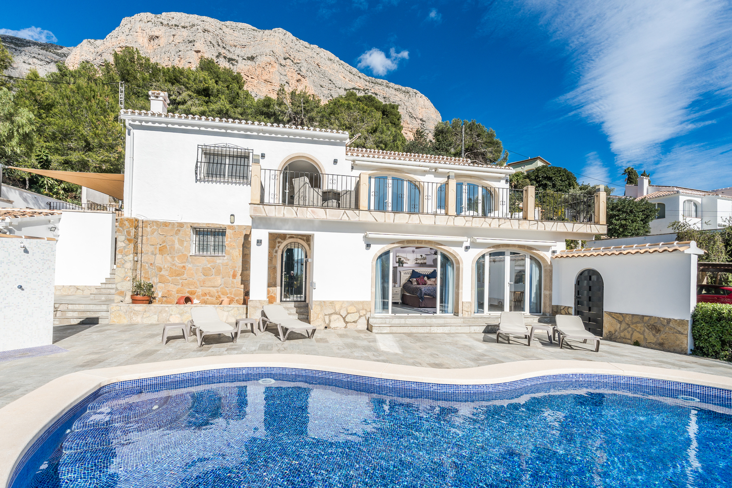 Villa exterior featuring a blue mosaic pool, stone-paved terrace with loungers, and traditional white facade set against the Montgó mountain.