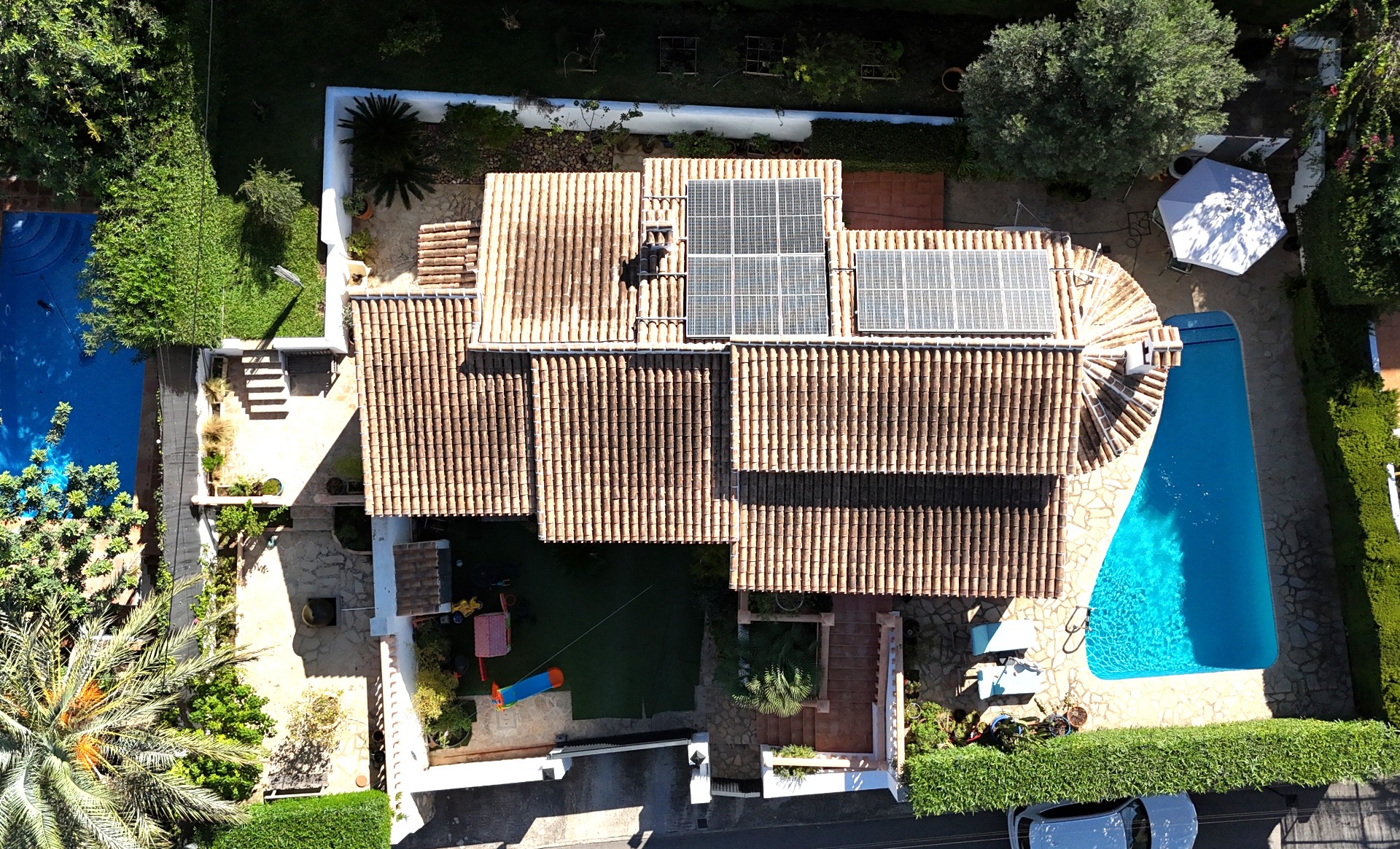 Aerial perspective of the villa featuring terracotta roof tiles with solar panels, a private swimming pool, and a Mediterranean garden with a children's play zone.