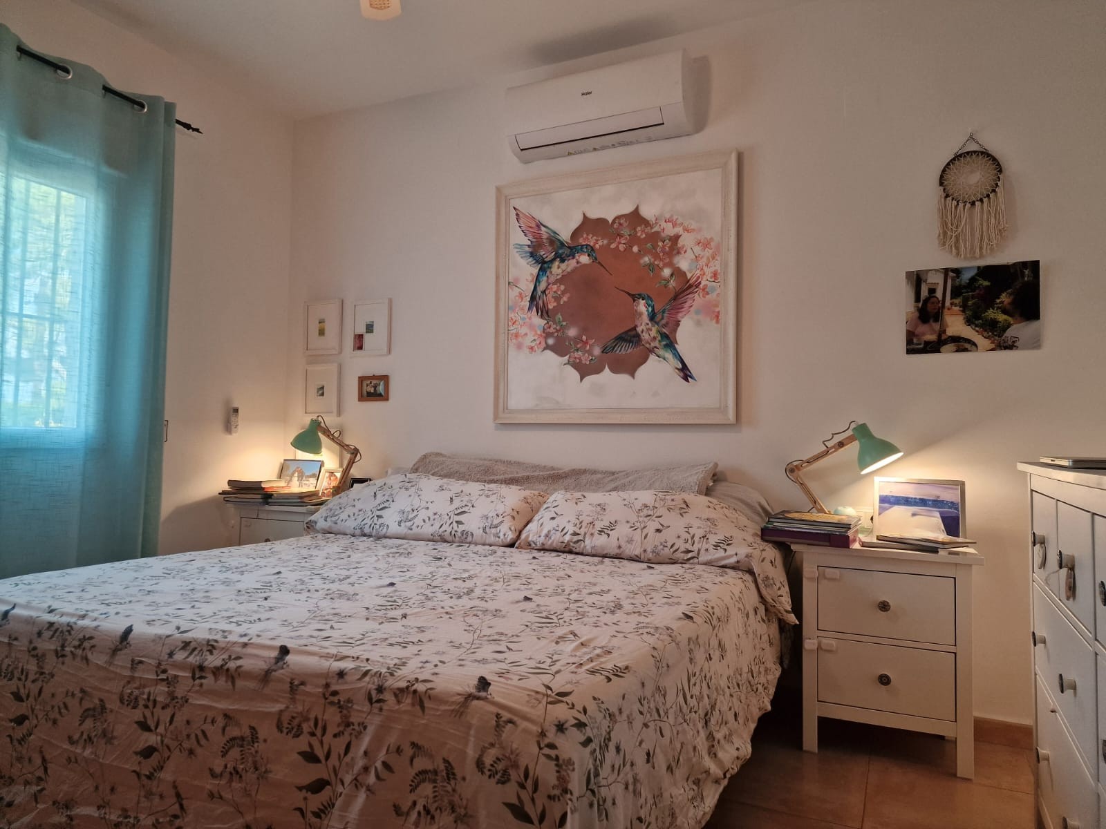 Bright bedroom featuring a double bed with botanical linens, Haier air conditioning, dual bedside tables with green lamps, and a large hummingbird artwork.