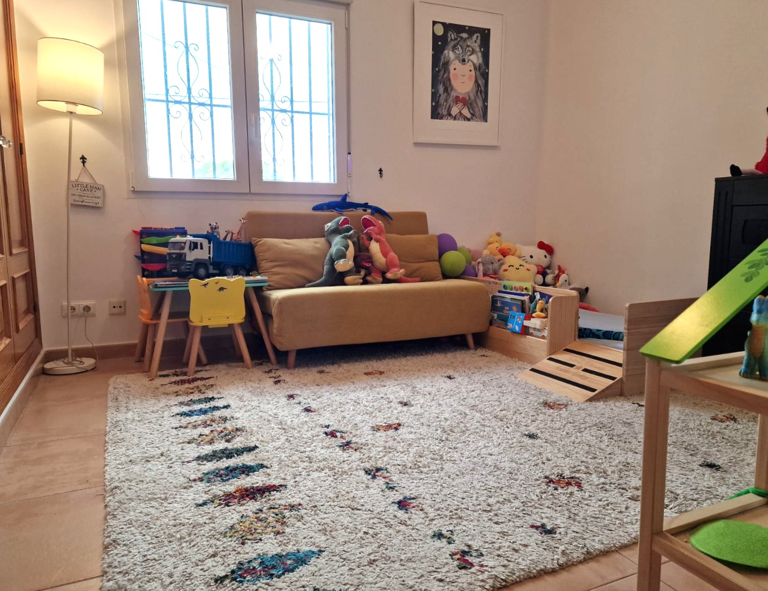 Dedicated children's play area featuring a textured rug, a yellow sofa, and a small activity table. The space includes natural light from a window with security bars.