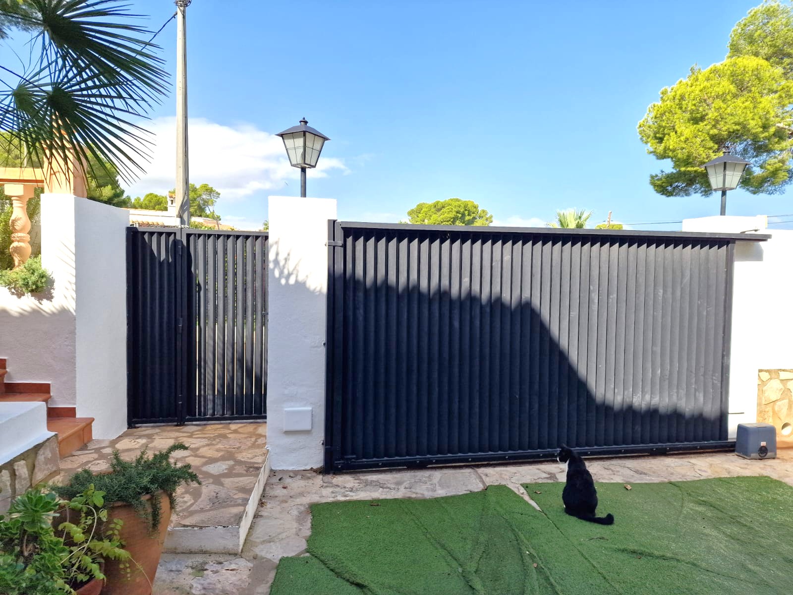 Entrance featuring a black sliding vehicle gate and matching pedestrian door, set between white pillars with classic lanterns and a stone-paved path.