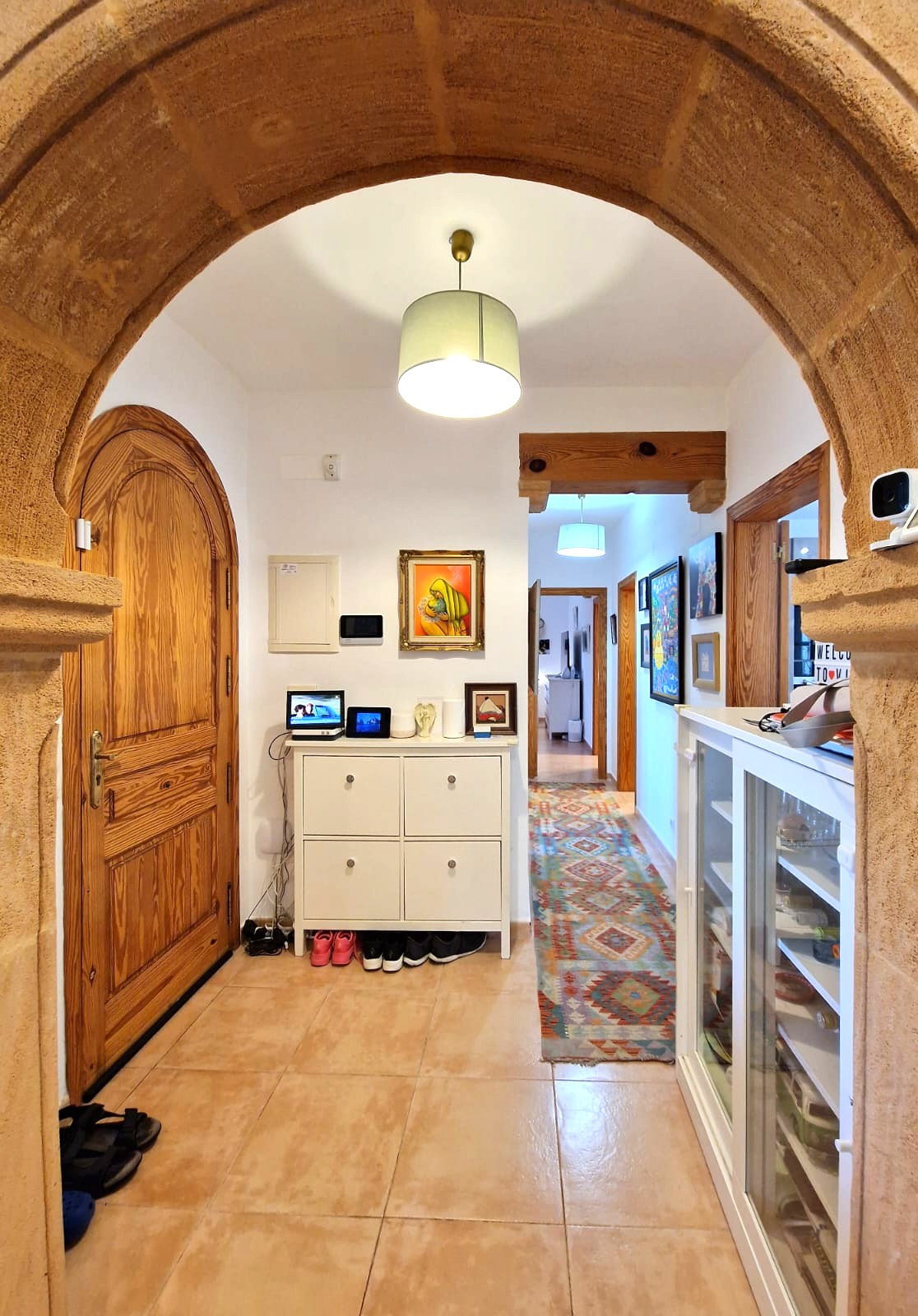 Entrance hall viewed through a traditional Tosca stone arch, featuring an arched wooden door, tiled flooring, and a hallway with a patterned runner and exposed timber beams.