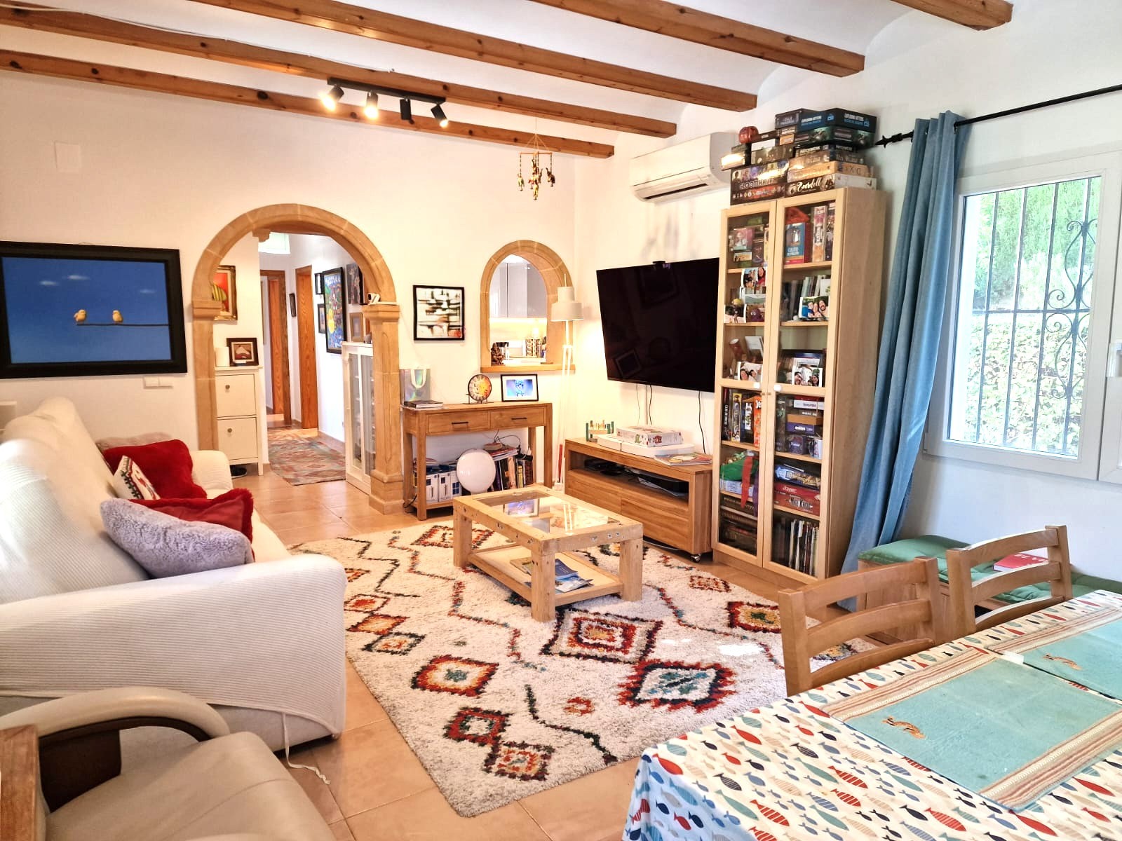 Living and dining area featuring exposed wooden ceiling beams, Tosca stone arches, and wall-mounted air conditioning. The space connects to the kitchen and hallway.