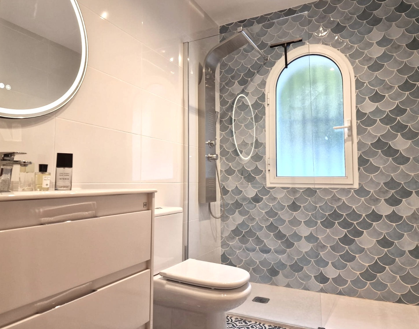 Modern bathroom featuring a walk-in shower with blue fish-scale tiles, an arched frosted window, a stainless steel shower column, and a backlit circular mirror.