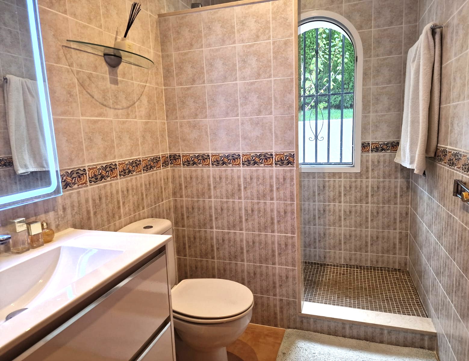 Modern bathroom featuring a walk-in shower with mosaic flooring, beige tiled walls with decorative borders, and an arched window providing natural light.