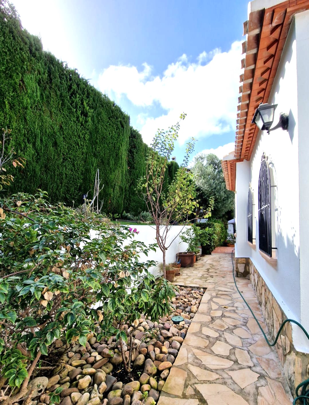Side walkway featuring natural stone paving, a white facade with stone accents, and high cypress hedges. Wrought iron window grilles and terracotta tiles add traditional character.