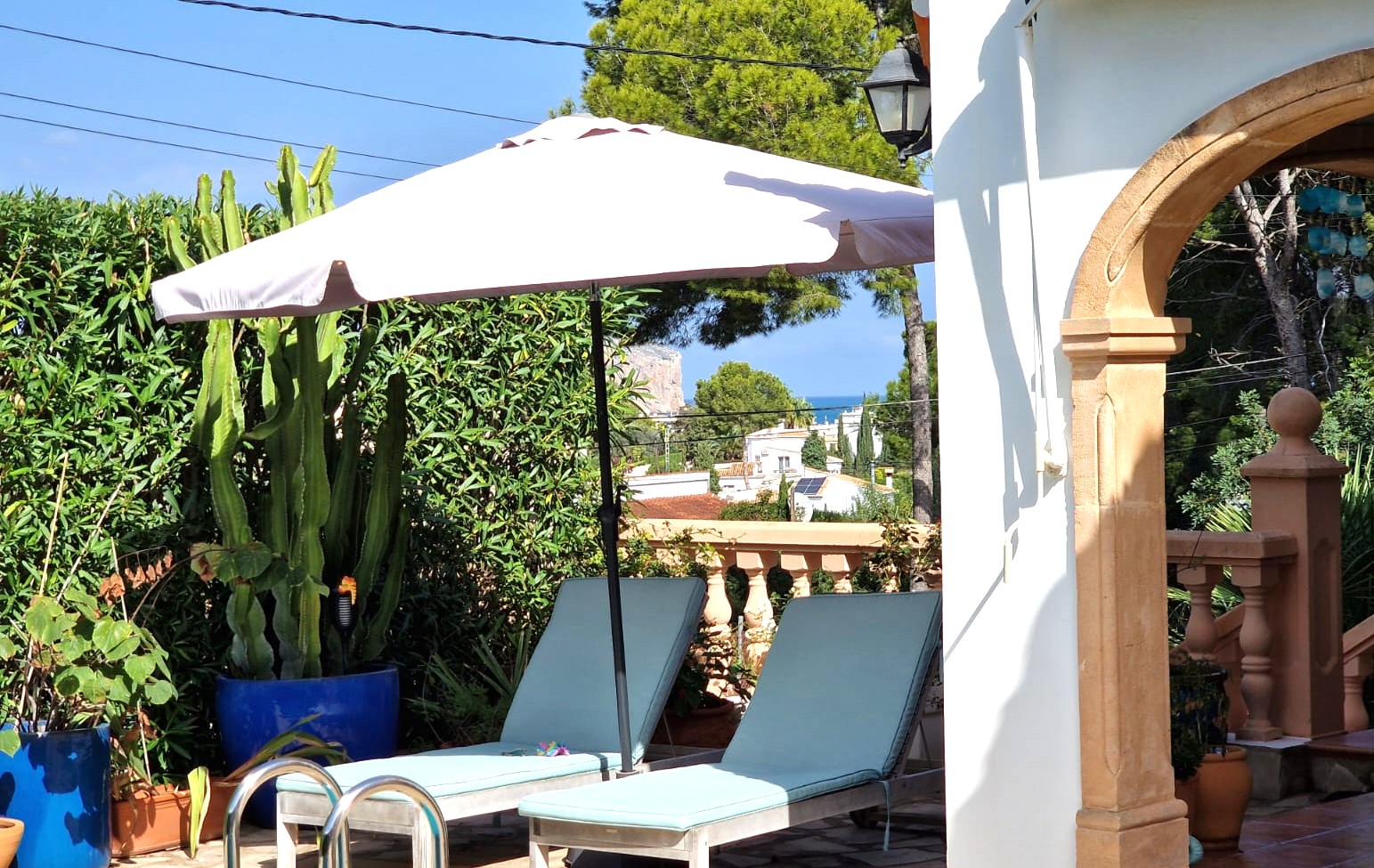 Terrace area featuring sun loungers under a parasol, a stone archway, and Mediterranean plants, offering a glimpse of the sea and Cabo San Antonio in the distance.
