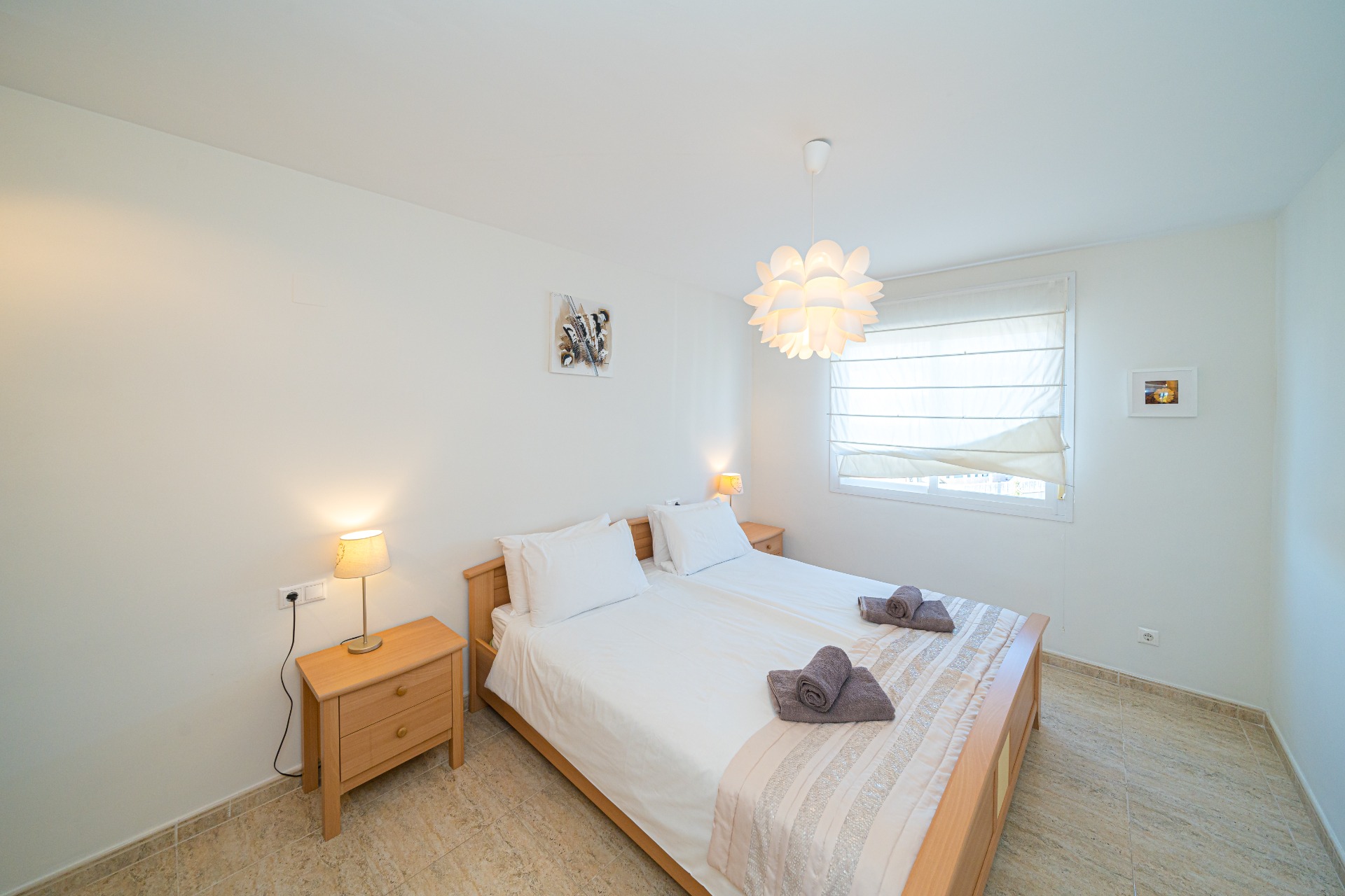 Bright bedroom featuring light wood furniture, a double bed, and matching nightstands. Natural light enters through a window with a white blind, highlighting the tiled flooring.