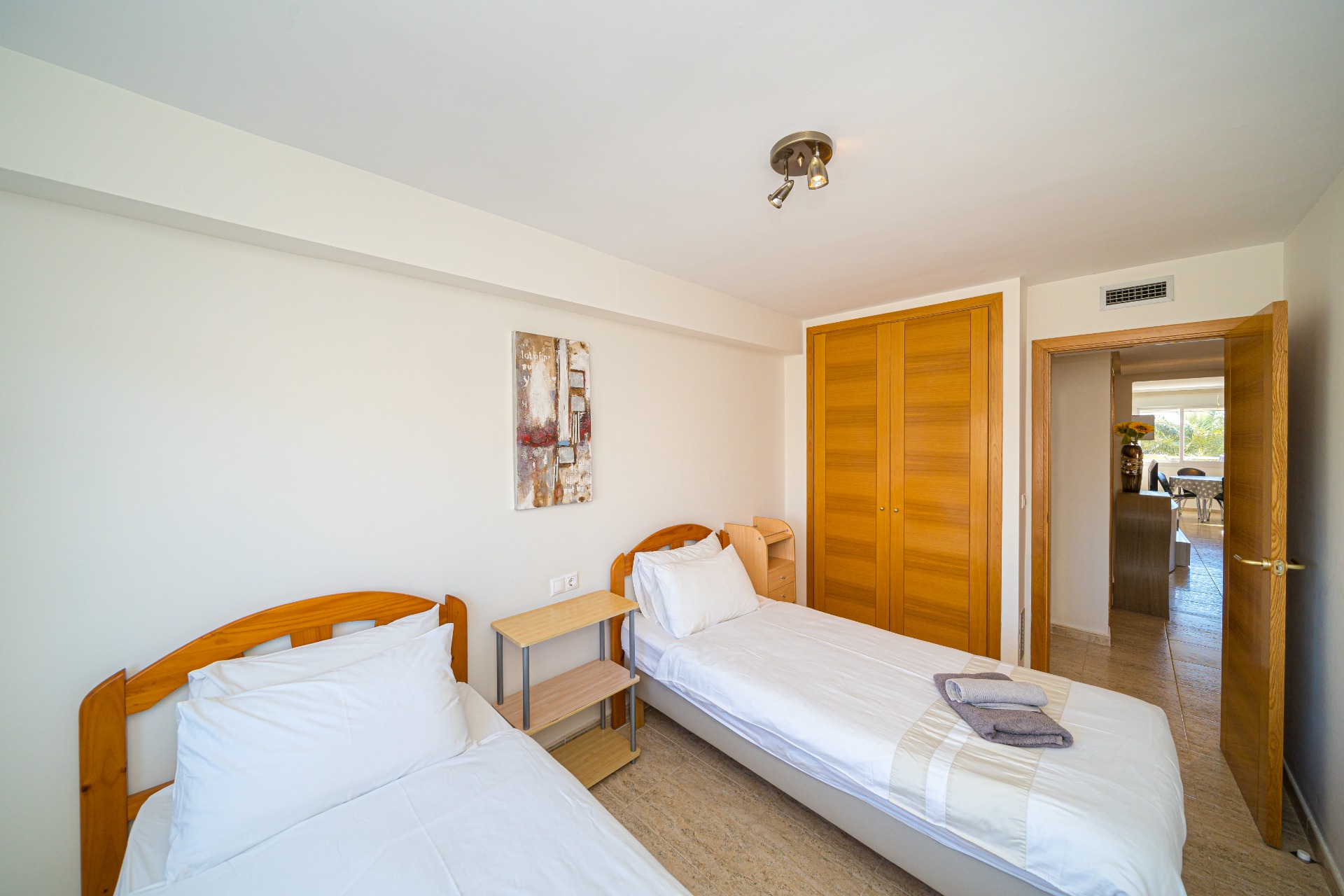 Bright bedroom featuring twin beds with wooden headboards, a built-in oak-finish wardrobe, and tiled flooring. An open doorway leads toward the living area.