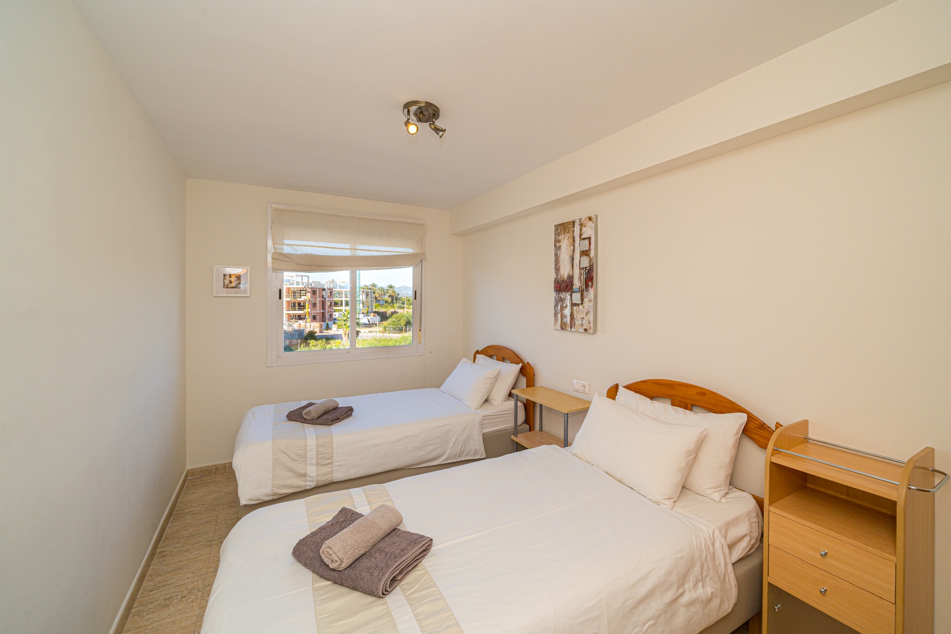 Bright bedroom featuring twin beds with wooden headboards, neutral tiled flooring, and a window providing natural light and views of the surrounding area.