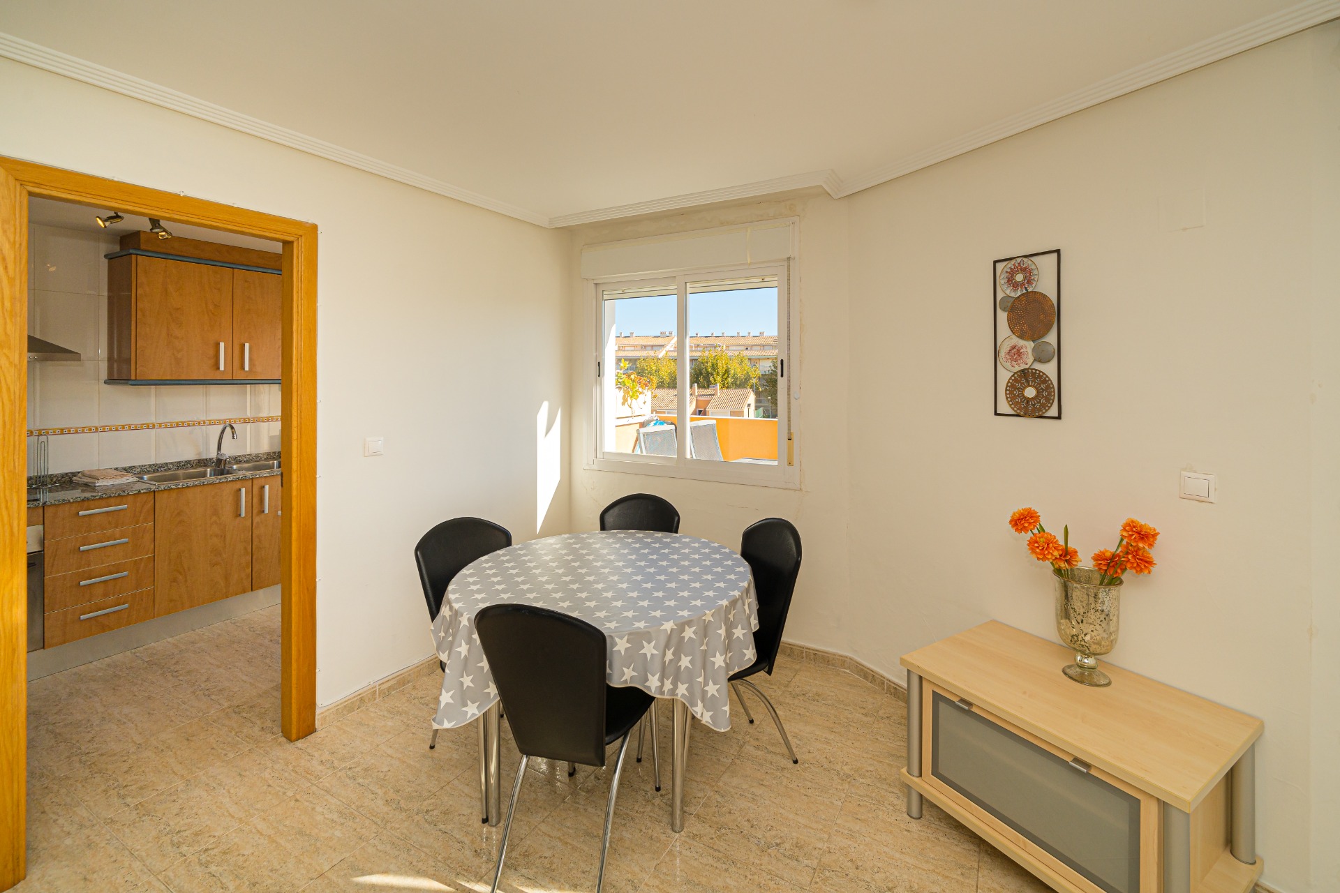 Bright dining area with a round table and tiled floors, offering a view through the window. The adjacent kitchen features wooden cabinetry and granite countertops.