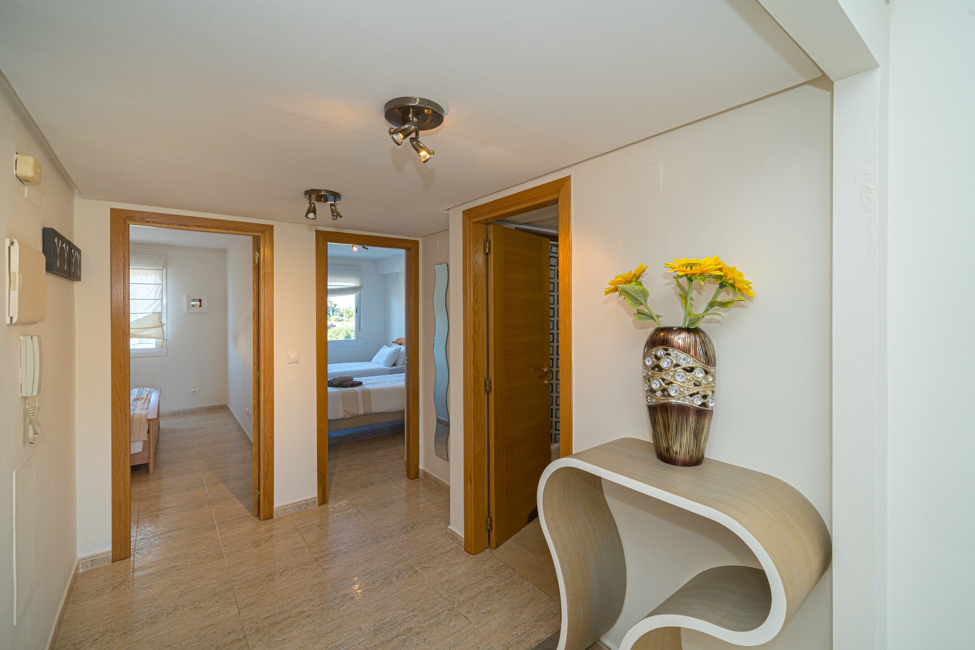 Central hallway with polished tile flooring leading to two bedrooms and a bathroom, featuring modern ceiling spotlights and a curved wood-effect console table.