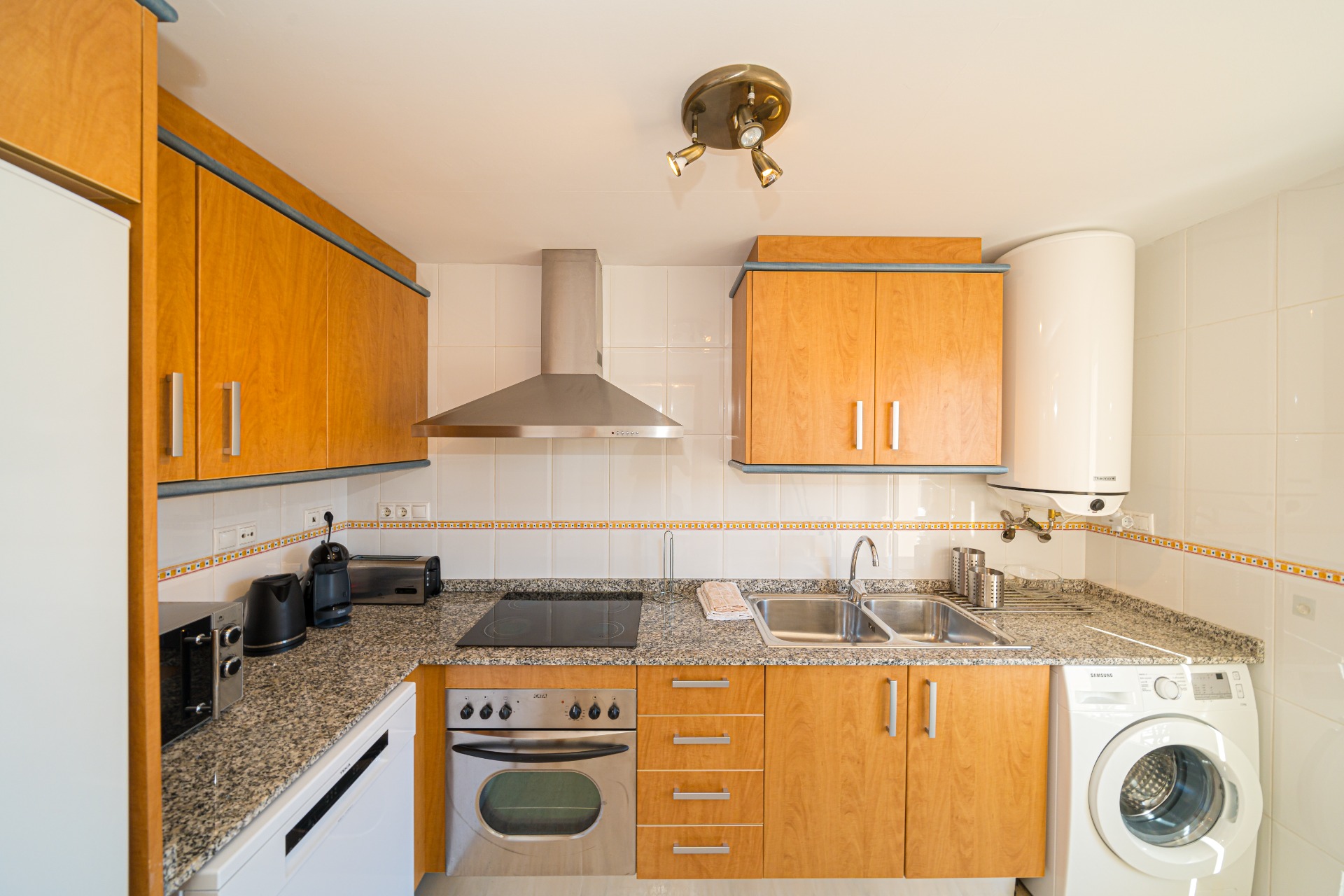 Fully equipped kitchen featuring wood-finish cabinetry, granite countertops, and integrated appliances including an oven, washing machine, and stainless steel extractor hood.