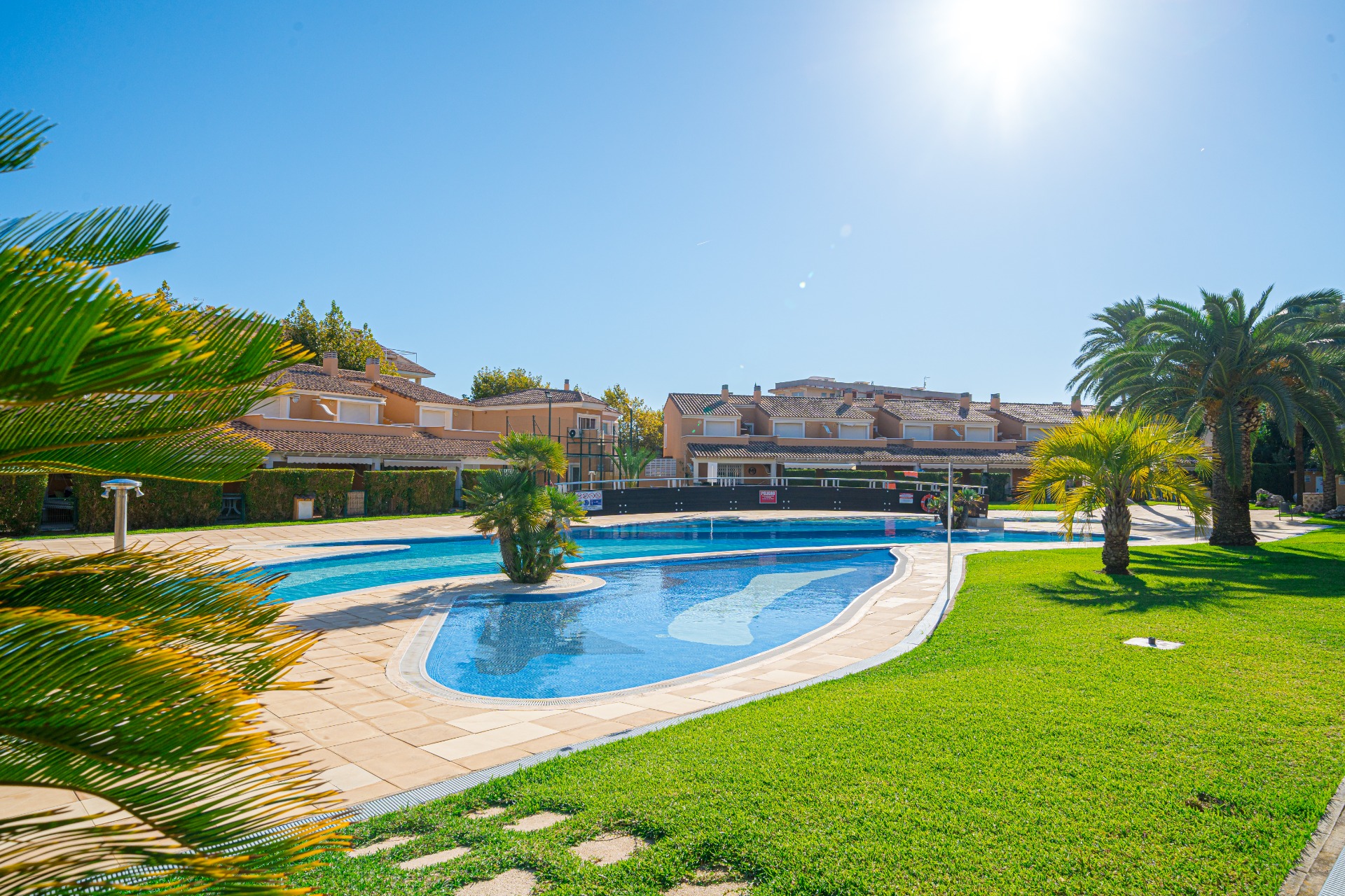 Large communal swimming pool featuring a central island and bridge, surrounded by manicured lawns and Mediterranean palms under a clear sky.