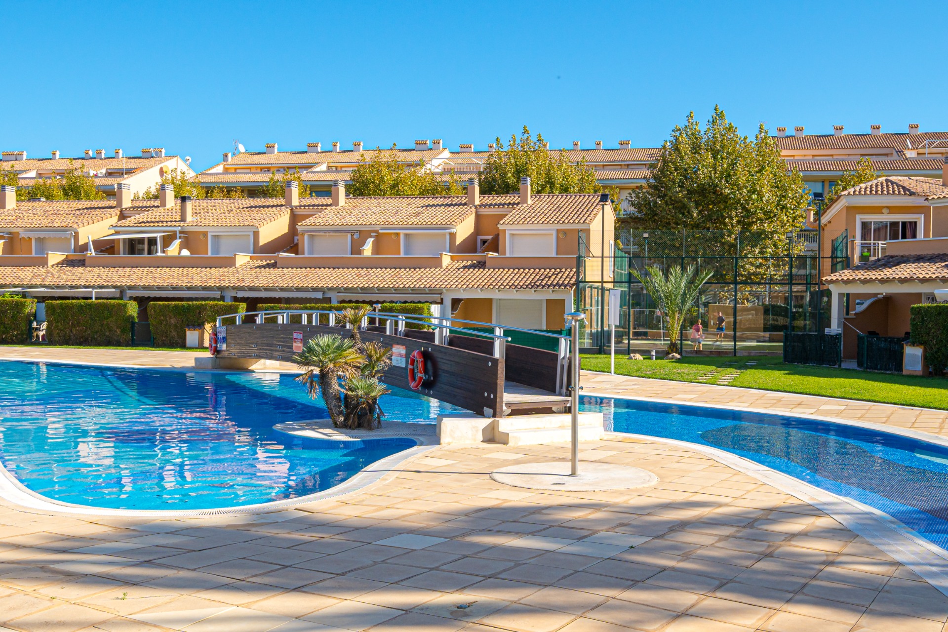 Large communal swimming pool featuring a wooden bridge, surrounded by a tiled sun terrace and a professional padel court in the background.