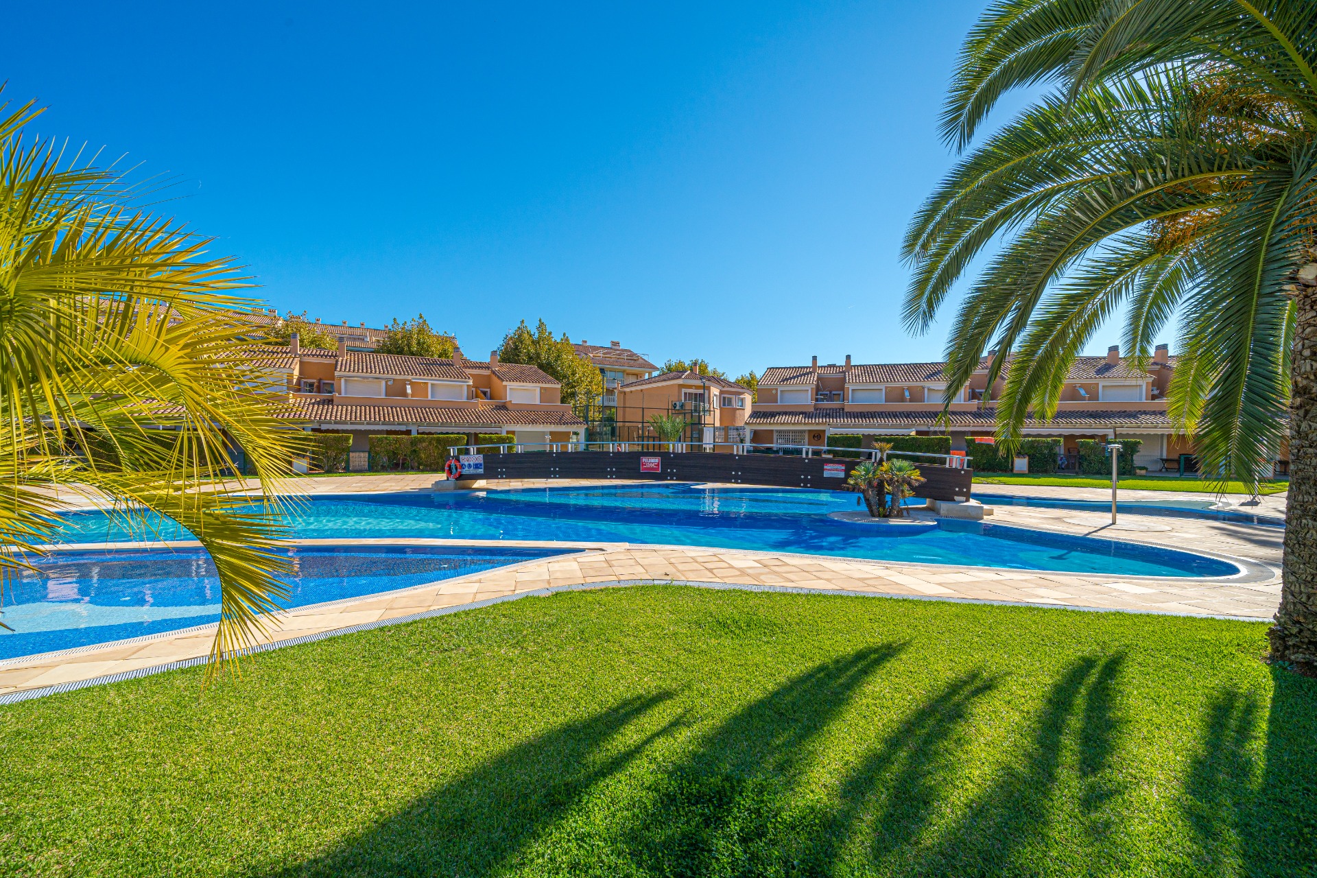Large communal swimming pool featuring a wooden bridge, surrounded by manicured lawns and palm trees in Moreras del Saladar.