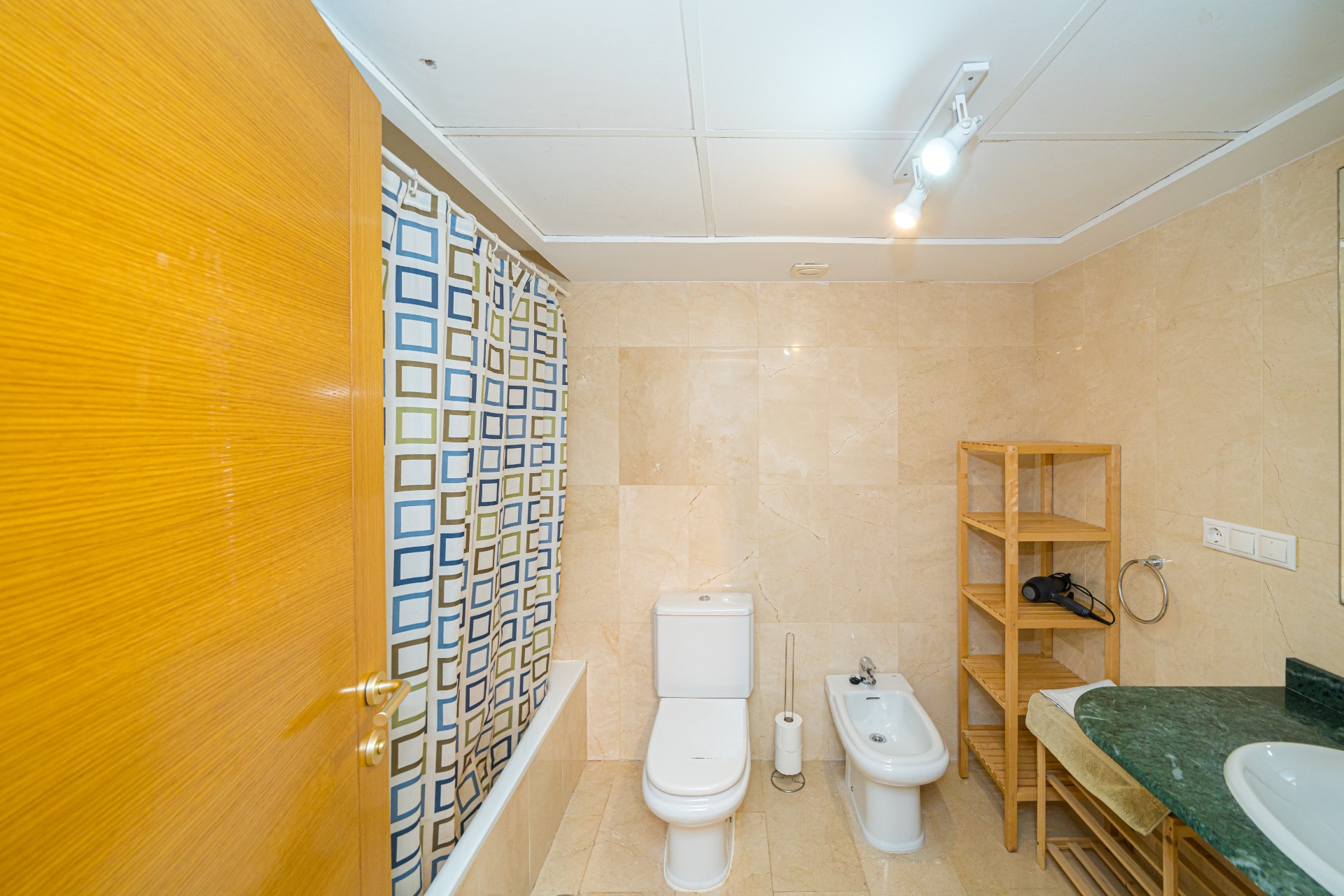 Modern bathroom featuring beige marble-effect tiling, a bathtub with a geometric curtain, a bidet, and a green marble vanity top with wooden shelving.