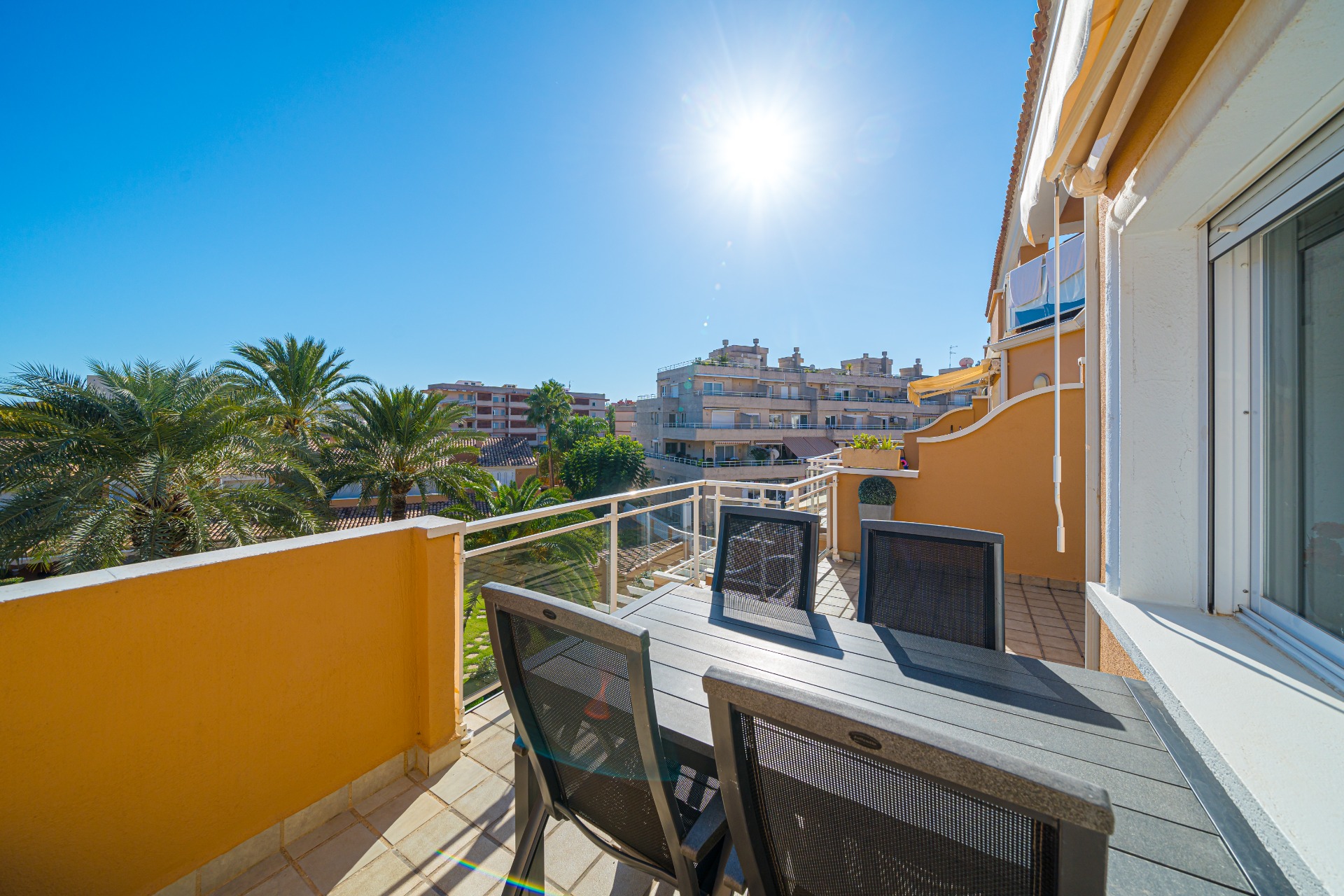Private sunny terrace with a dark grey outdoor dining set, tiled flooring, and views overlooking palm trees and neighboring residential complexes.