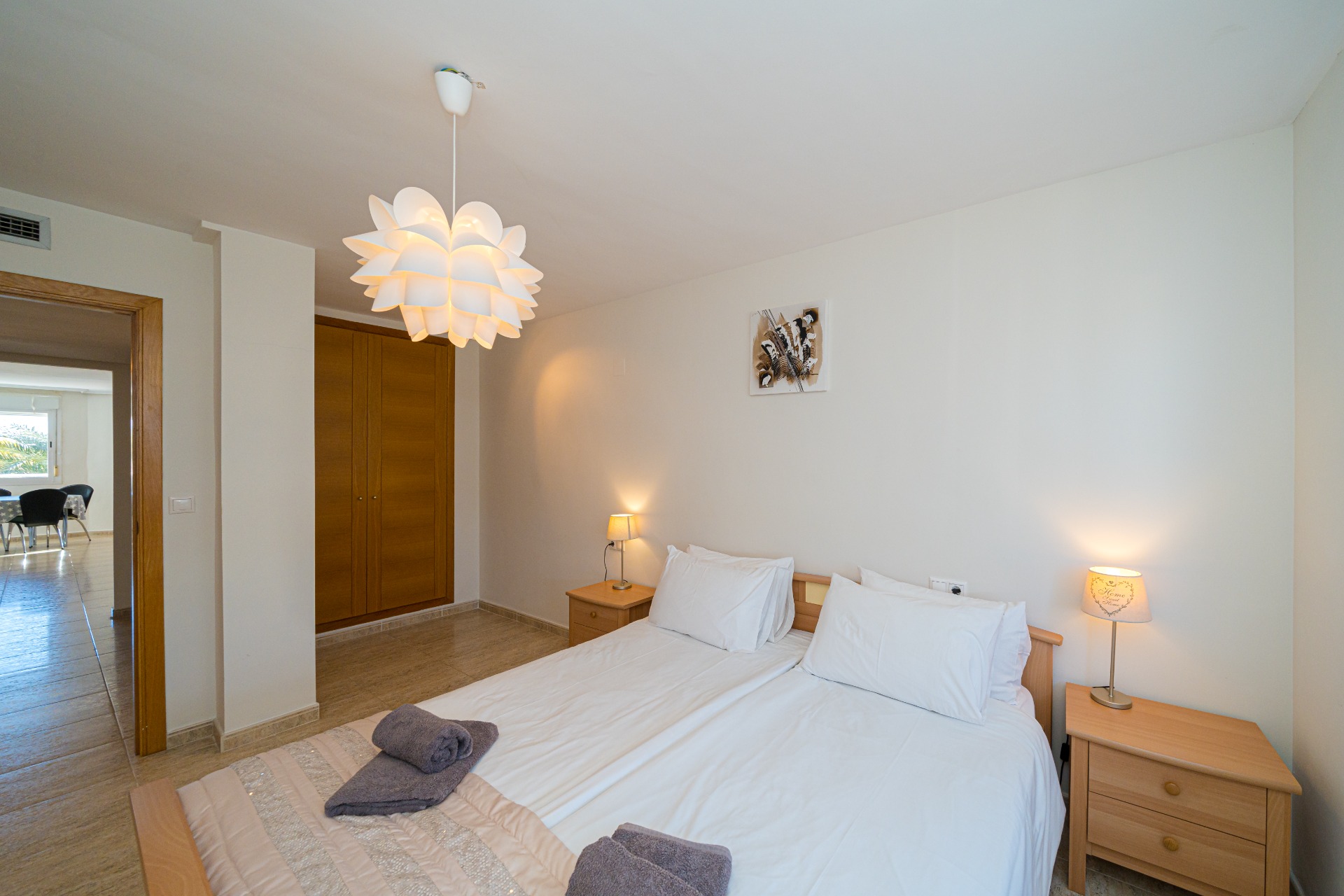 Spacious bedroom featuring twin beds, built-in wooden wardrobes, and a decorative lotus-style pendant light. The doorway reveals a bright dining area beyond.