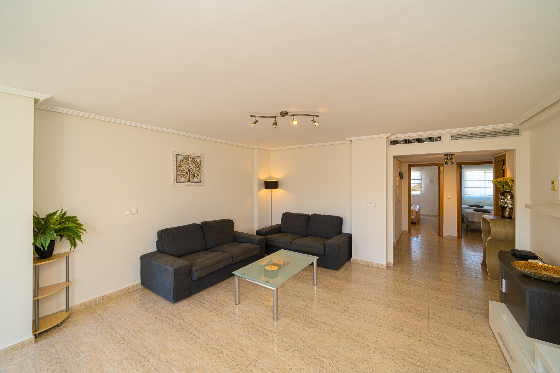 Spacious living room featuring dark grey sofas, a glass coffee table, and polished tile flooring. The open layout leads to a hallway connecting the bedrooms.
