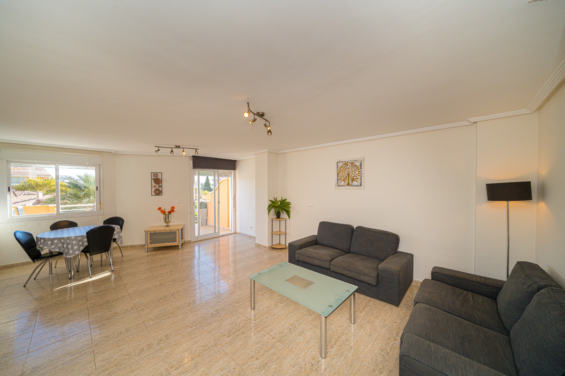 Spacious open-plan living area featuring polished tile flooring, dark grey sofas, and a dining set. Large sliding doors and windows provide ample natural light and terrace access.