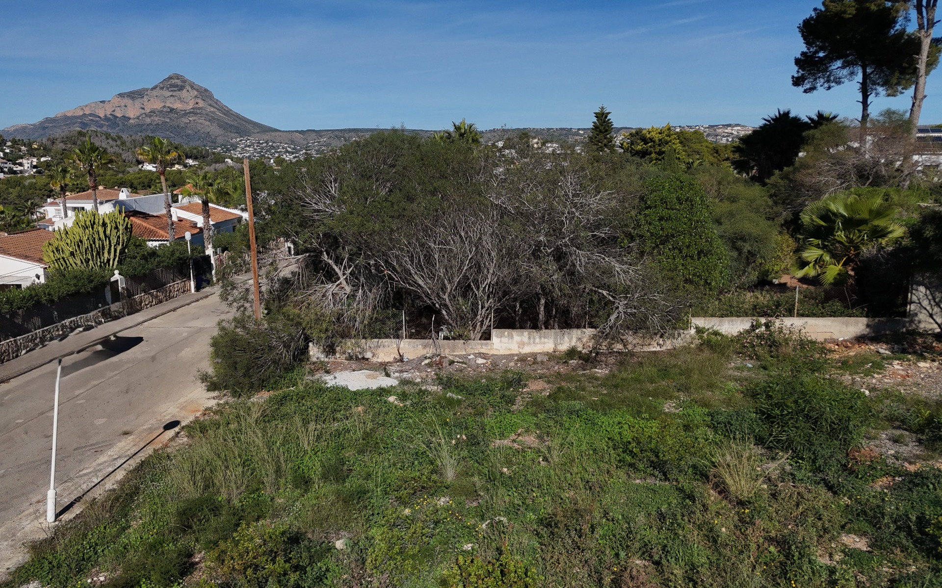 Elevated view of the flat 889 m² building plot in Pinosol, showing the quiet residential street, perimeter walling, and clear views toward the Montgó mountain.