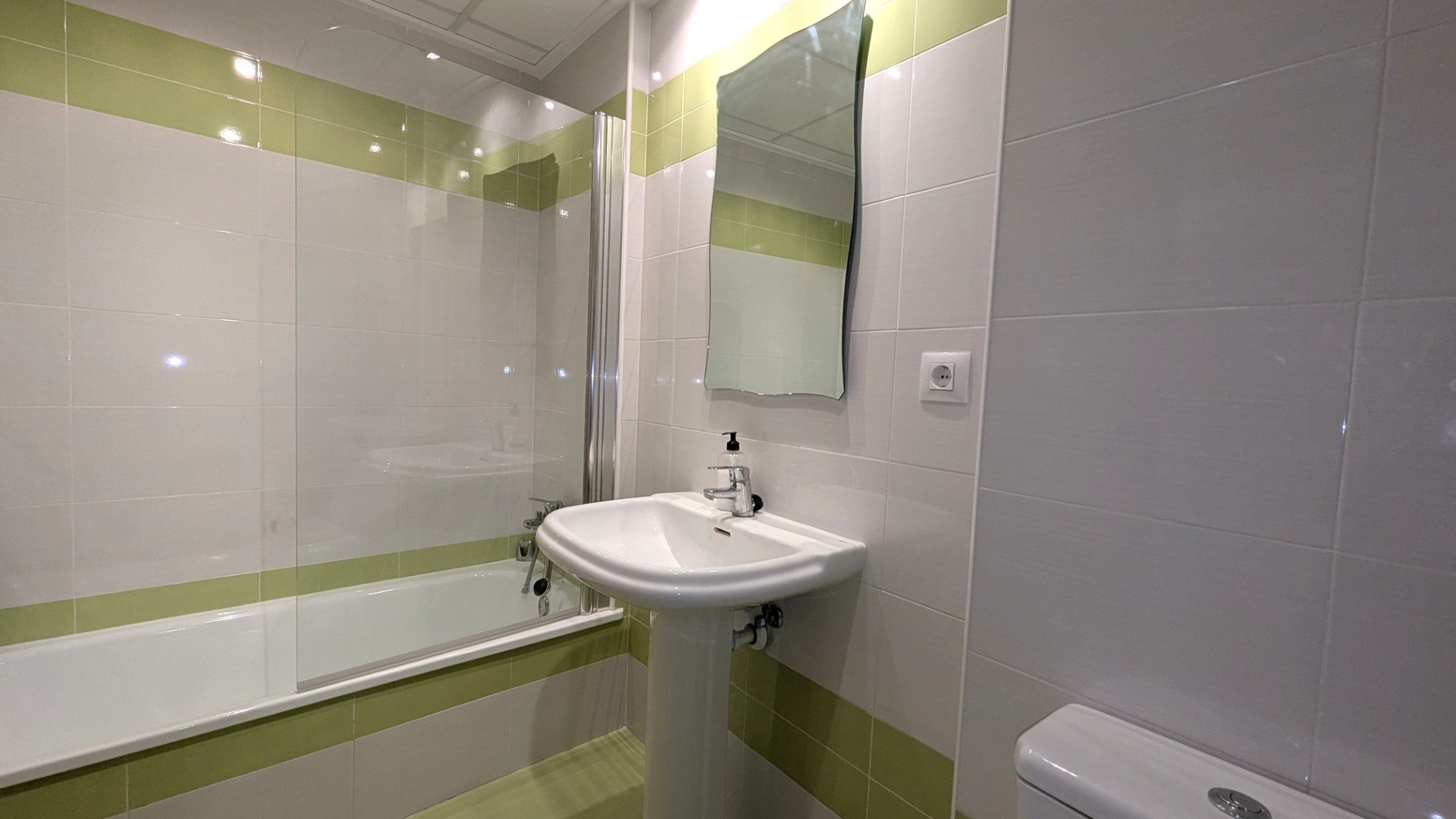 Bathroom featuring a bathtub with a glass partition, a pedestal sink, and a wavy mirror, accented by white and lime green horizontal tiling.