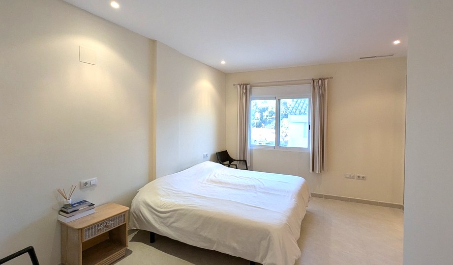Bright bedroom featuring neutral tones, recessed ceiling spotlights, and a large window with shutters. Includes tiled flooring and a wooden bedside table.