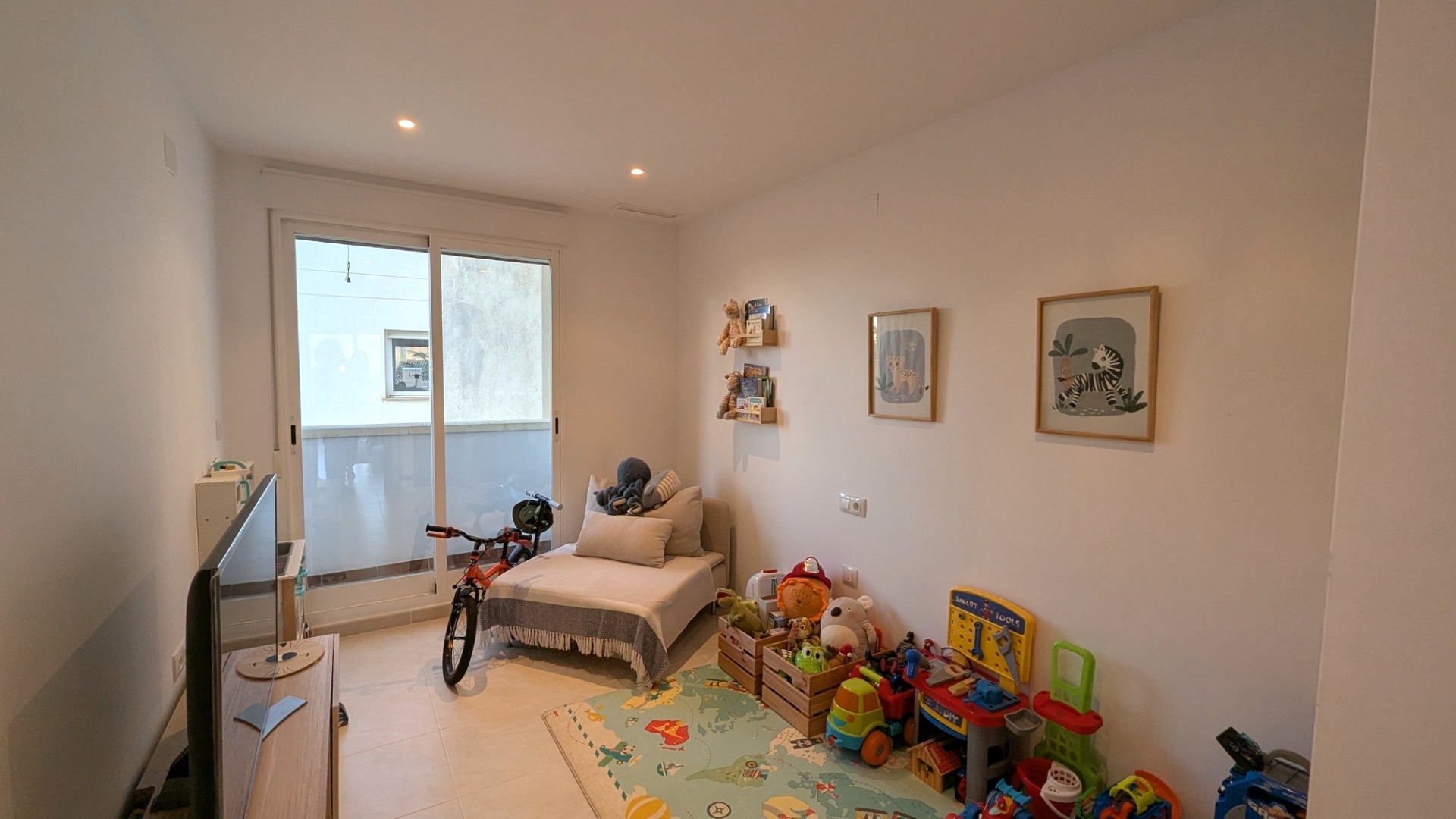 Bright bedroom used as a playroom, featuring recessed lighting, light tile floors, and sliding glass doors that open onto a private terrace.