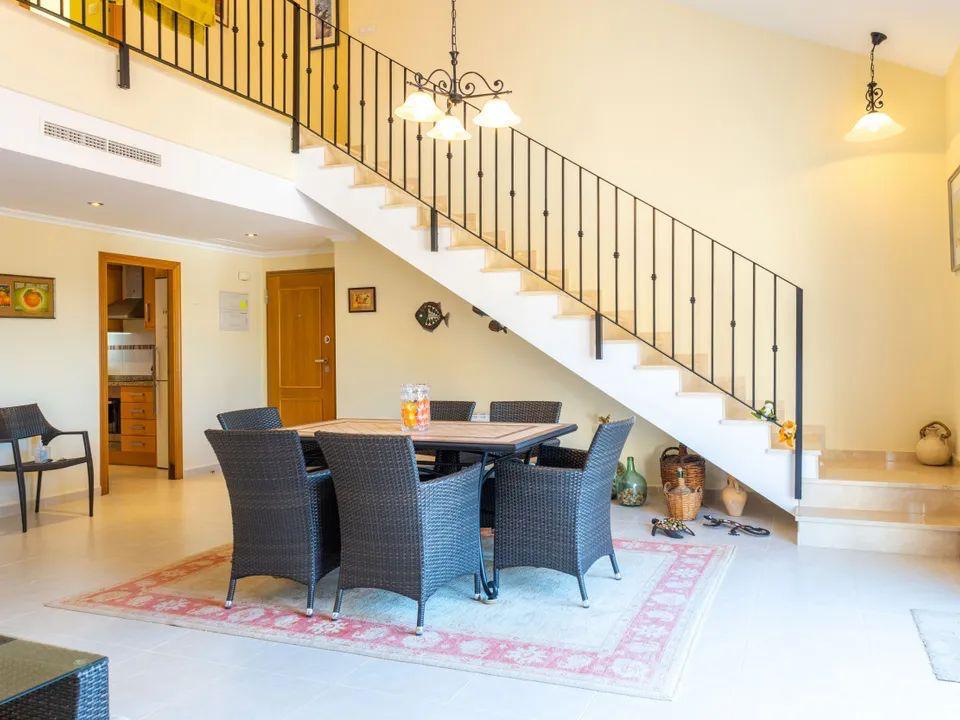 Dining area featuring a dark wicker set positioned under a light-colored staircase with wrought iron railings. Open-plan layout with tiled flooring and natural light.