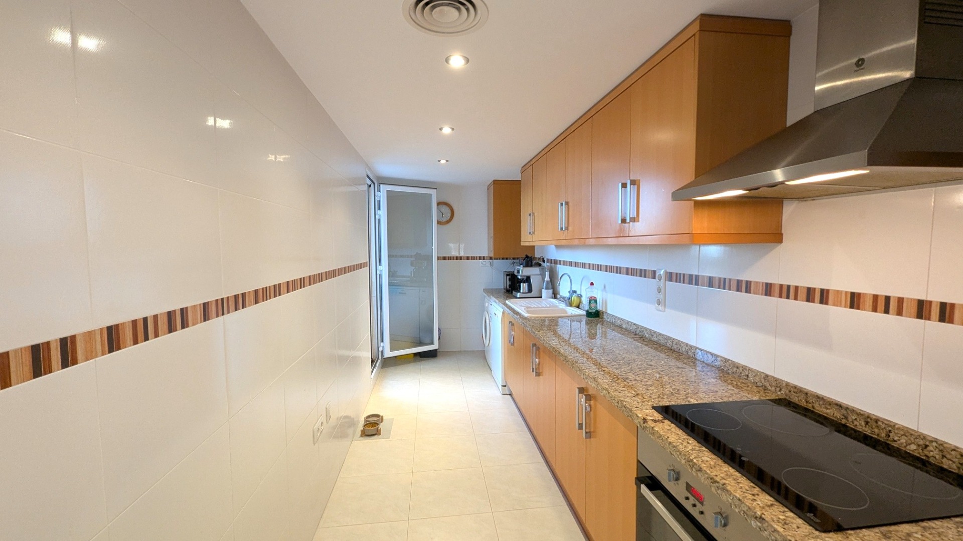 Independent kitchen featuring wood-finish cabinetry, granite countertops, and integrated appliances. The space includes white tiled walls with decorative borders and recessed lighting.