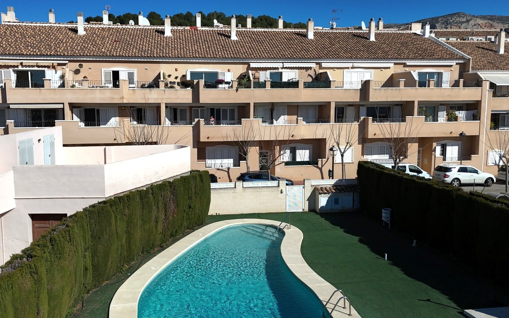 View of the communal kidney-shaped swimming pool surrounded by artificial turf and high privacy hedges, set against the residential facade with white shutters.