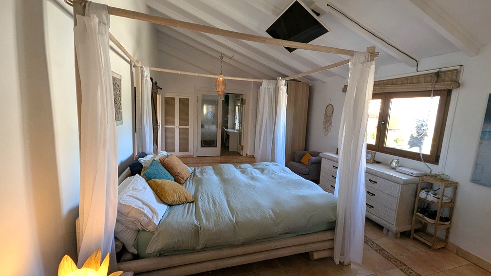 Bedroom featuring a bamboo four-poster bed, vaulted white-beamed ceilings, and terracotta tile flooring. Includes a ceiling-mounted TV and built-in storage.
