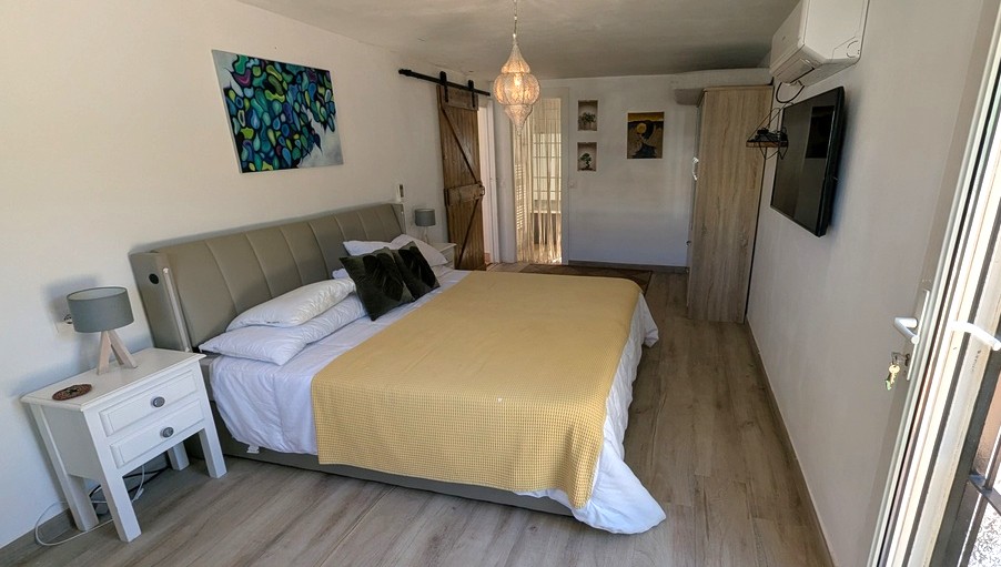 Bedroom featuring wooden flooring, a rustic sliding barn door, and a hanging lantern. Includes a wall-mounted TV, air conditioning, and a large bed with a grey headboard.