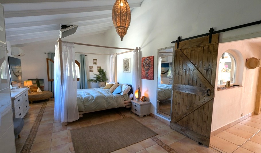 Bedroom with a four-poster bed, white beamed ceilings, and a rustic sliding barn door. Decorative terracotta tiles and a Moroccan lamp enhance the Mediterranean style.