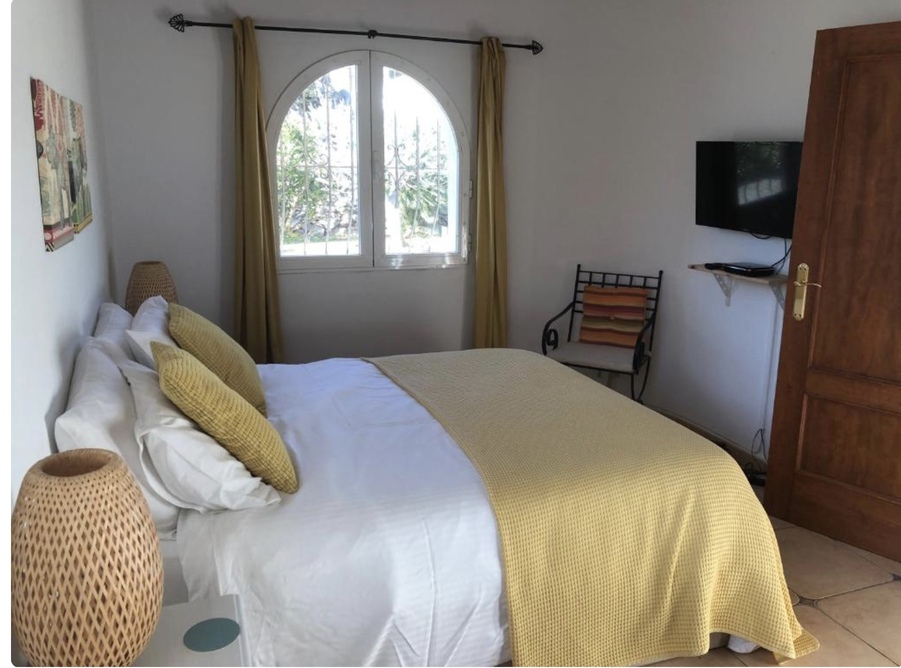 Bright bedroom featuring an arched window, terracotta flooring, and a wall-mounted TV. The space is decorated with mustard accents and woven bedside lamps.