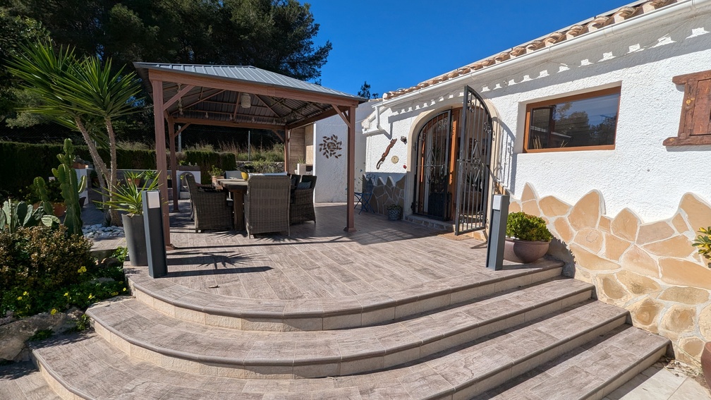 Elevated terrace with stone-effect tiling and curved steps leading to a shaded dining area under a wooden pergola, set against a white facade with decorative natural stone accents.